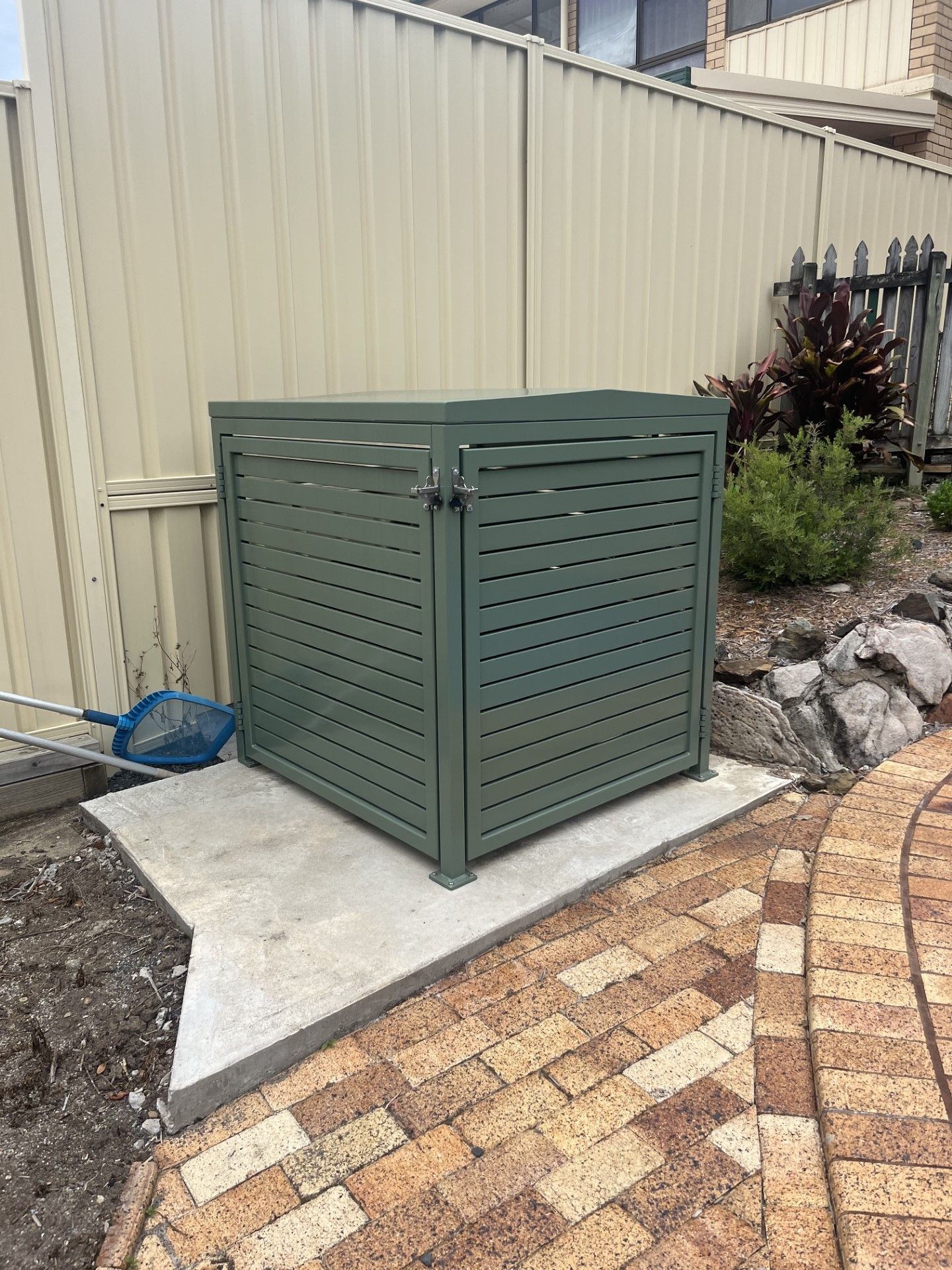 A green trash can is sitting on a brick sidewalk next to a fence — Fabricator In Coffs Harbour, NSW