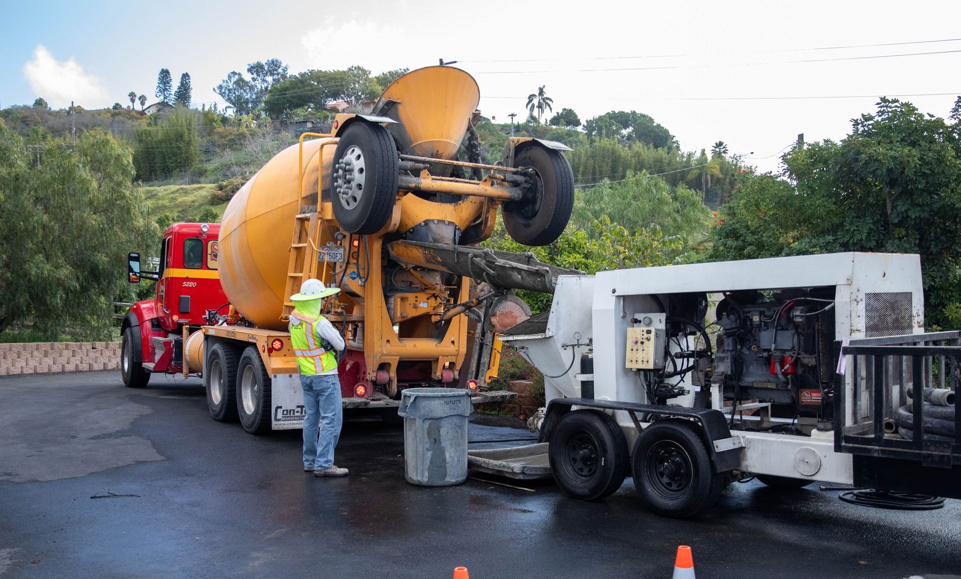 A worker in a safety vest stands by a concrete mixer truck pouring into a pump machine on a paved outdoor site.