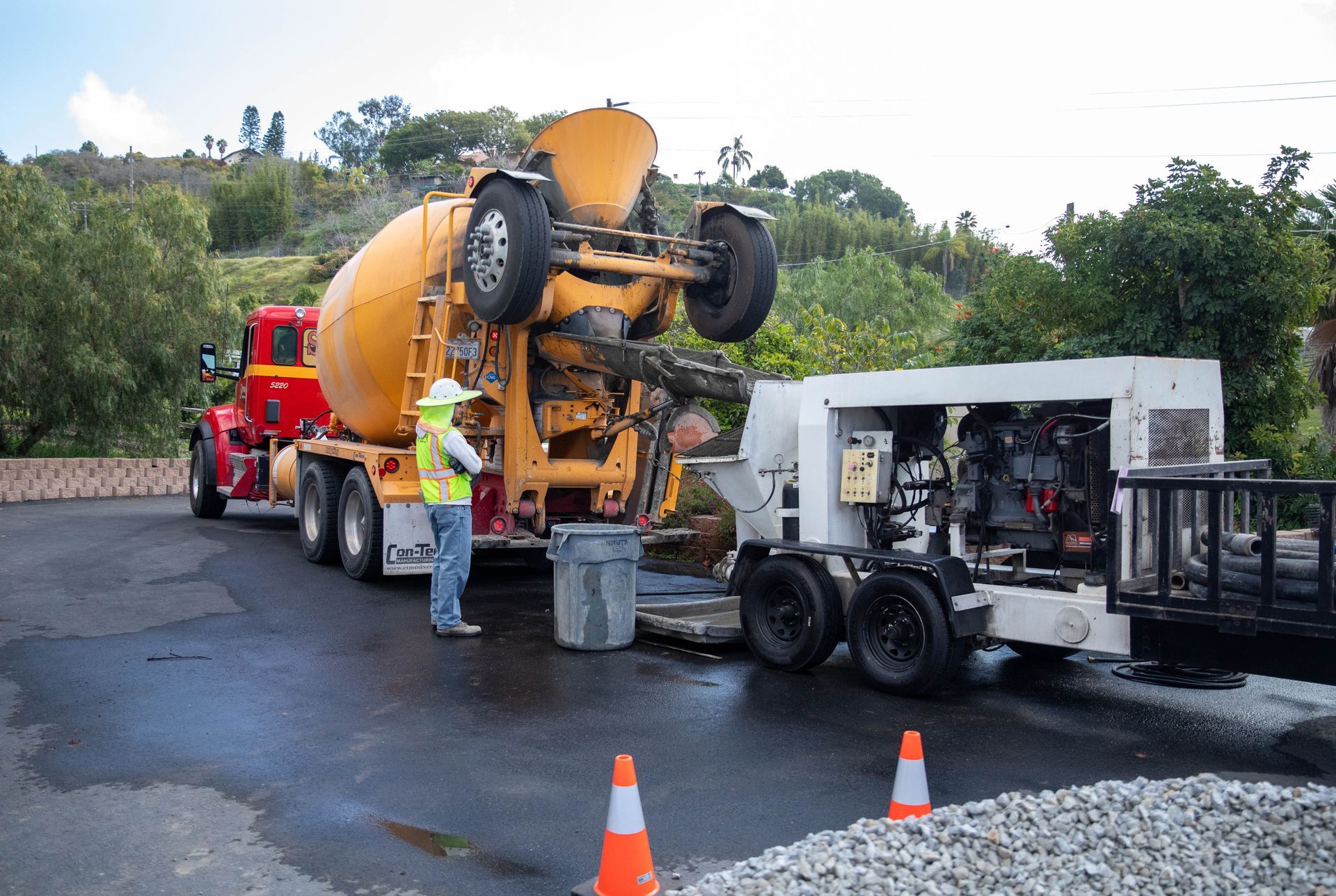 A construction worker in a high-visibility vest stands between a yellow cement truck and a white concrete pump.