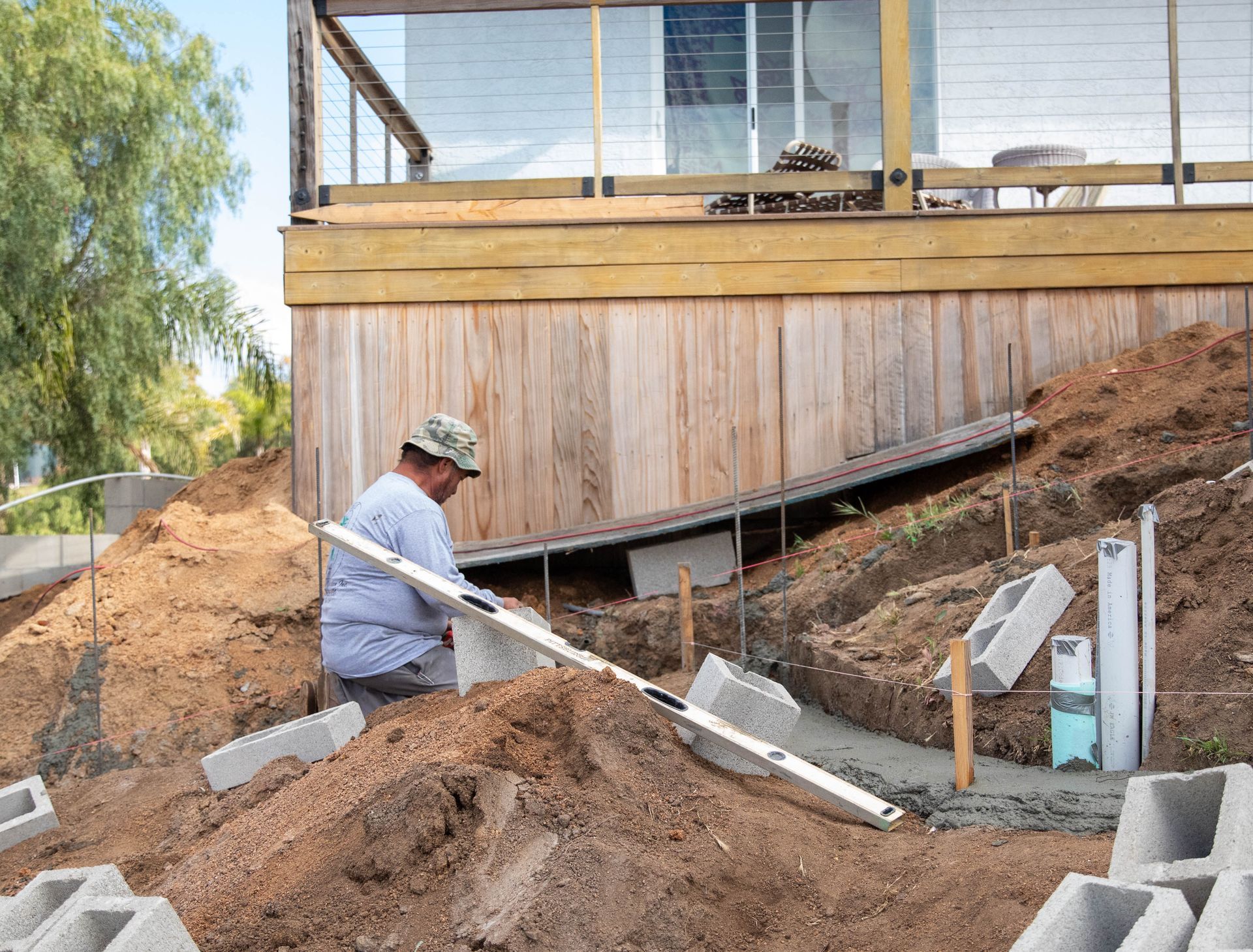 A construction worker levels concrete footings for a wooden deck addition on a sloped, excavated dirt site.