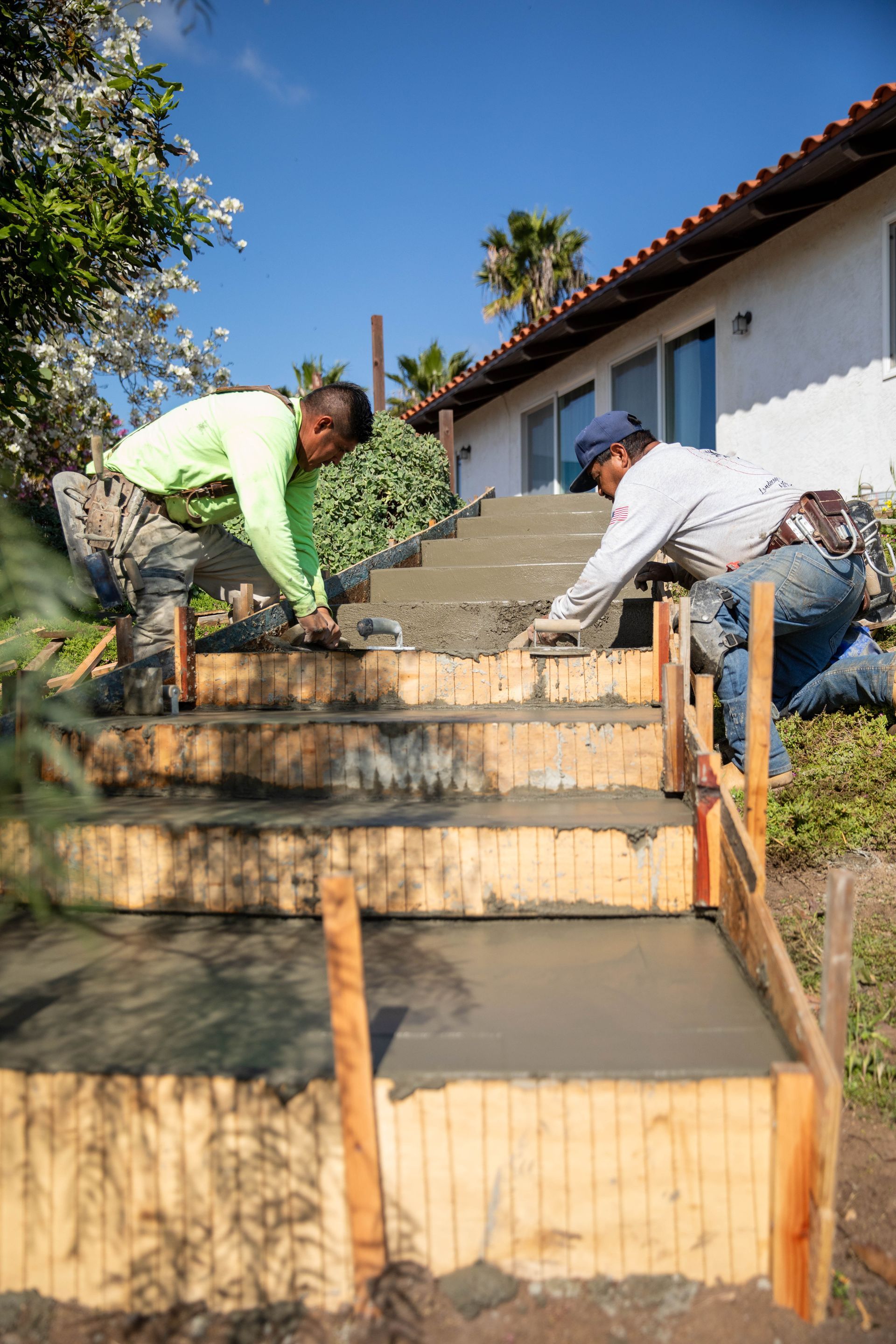 Two construction workers kneeling on wooden forms, smoothing fresh concrete on a set of outdoor residential stairs.