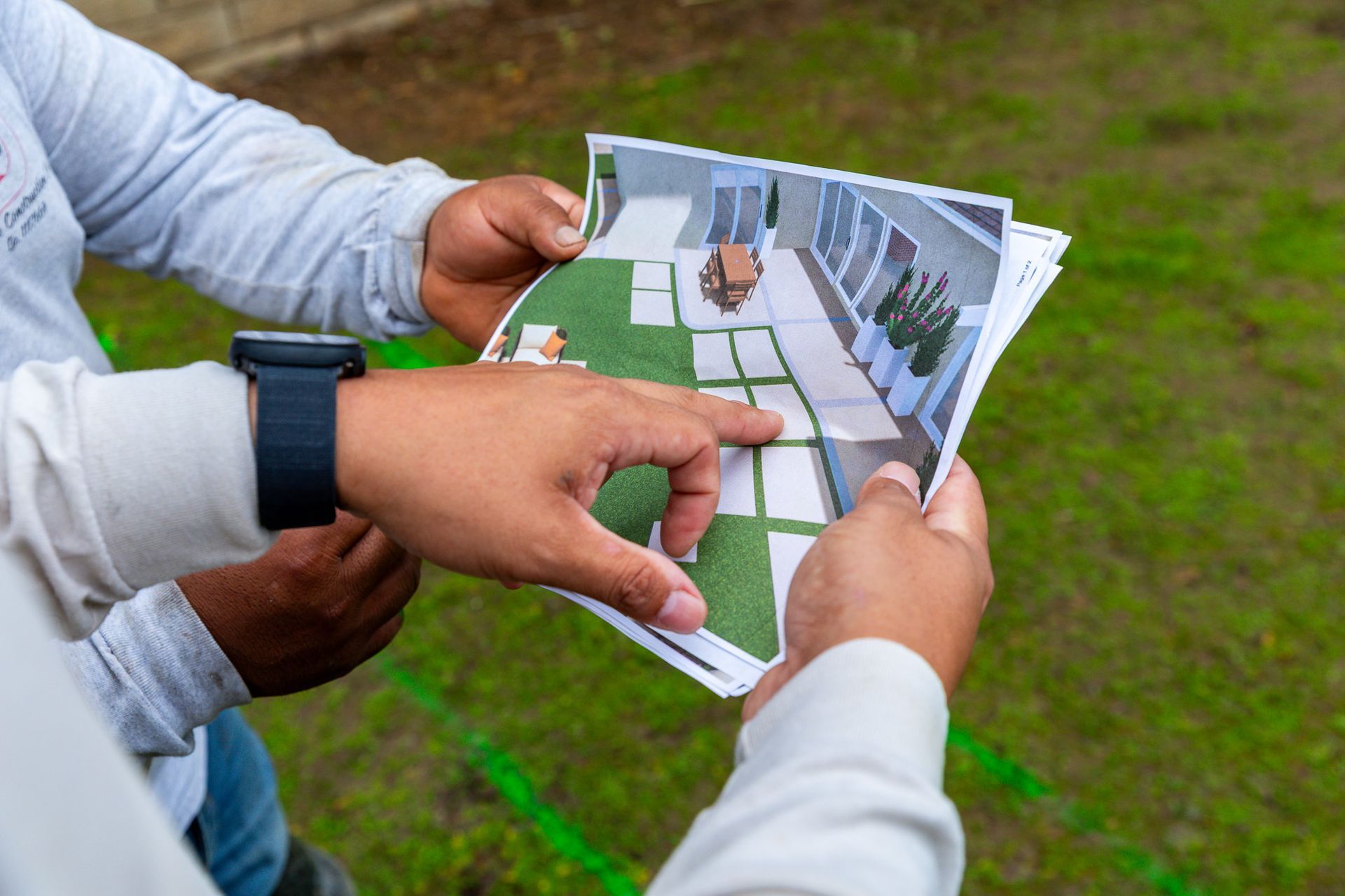 Two people examining a design plan outdoors, one pointing at the layout, on a green lawn.