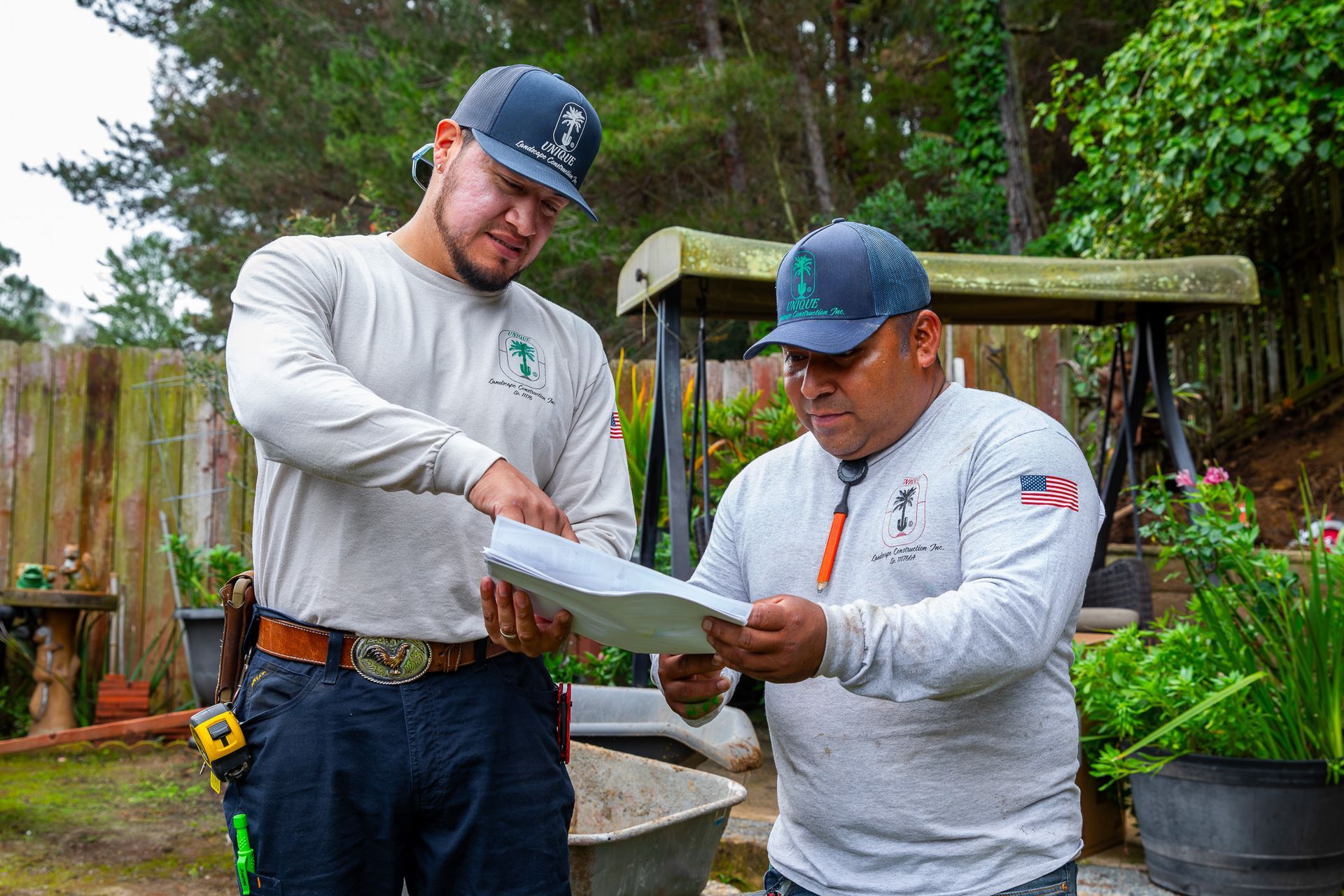 Two men in work shirts review plans outdoors near a fence and garden.