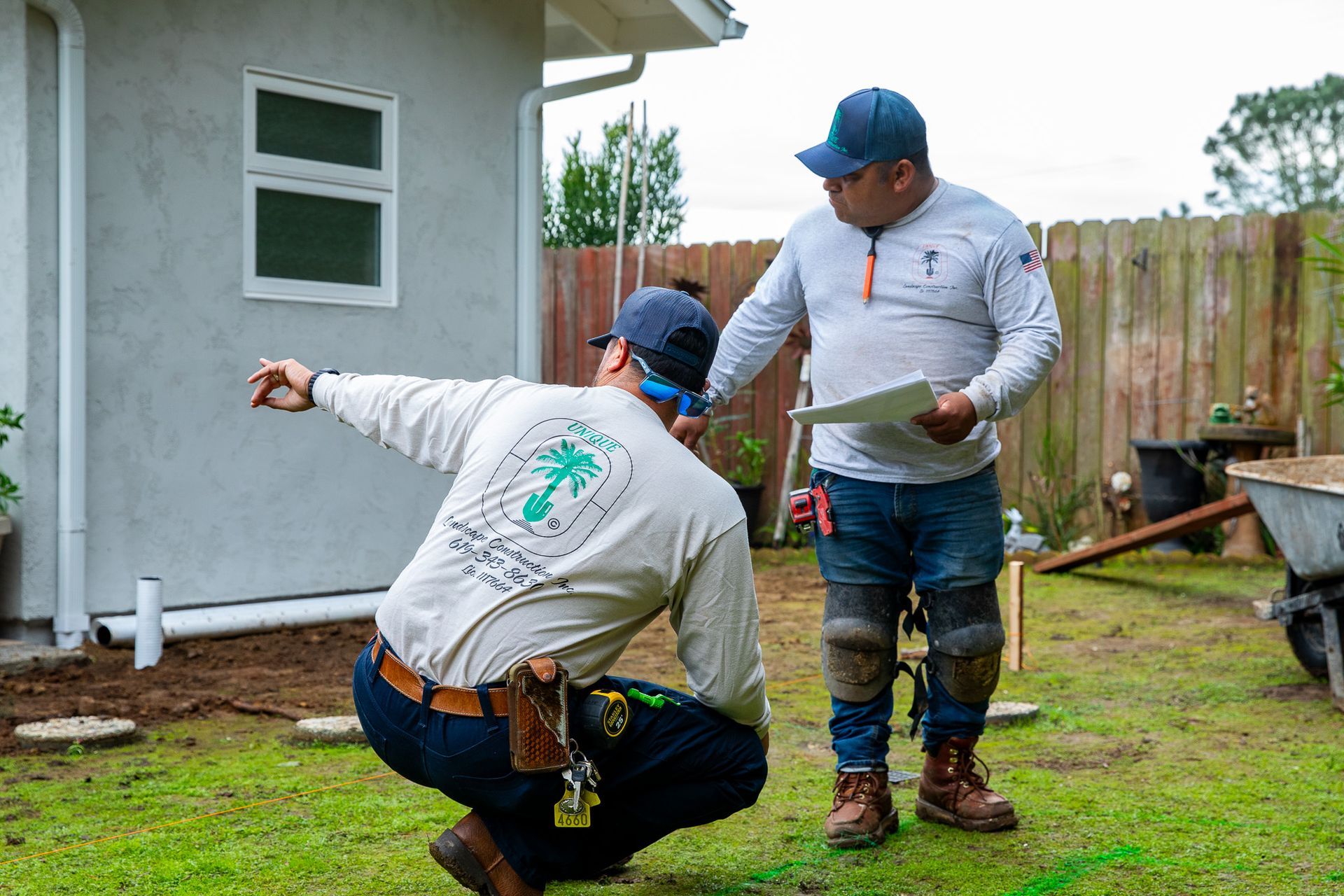 Two men examining a yard. One points, the other holds a paper. Near a house and fence.