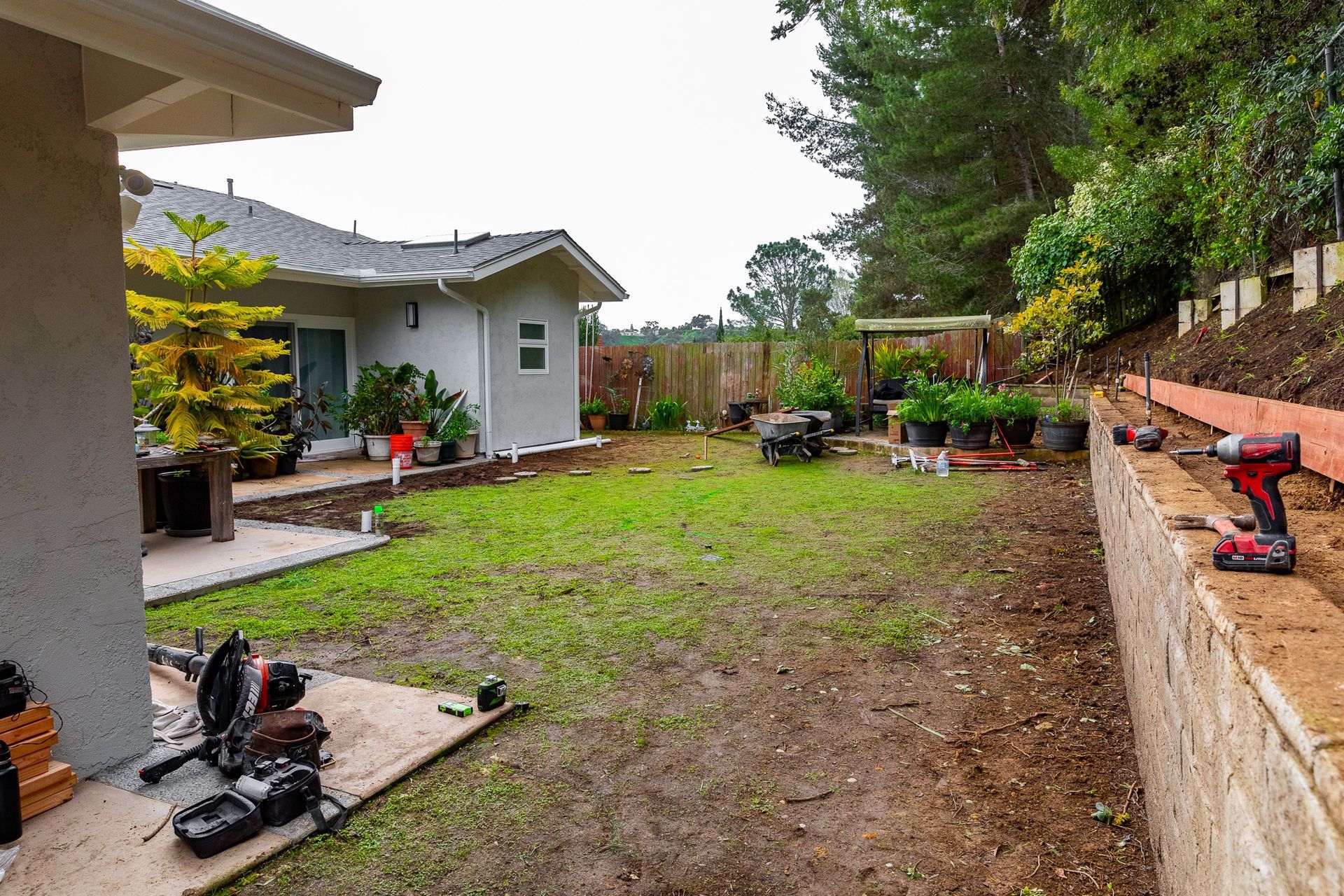 Backyard construction scene: grass, house, retaining wall with tools, trees, and gardening supplies.