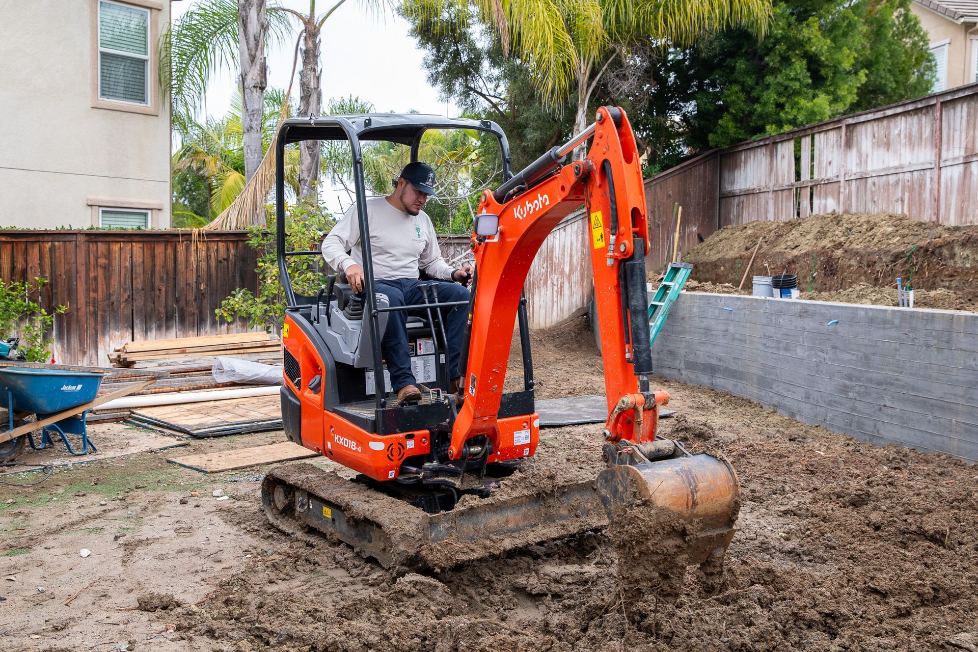 Man operating an orange Kubota excavator in a backyard, digging in the dirt.