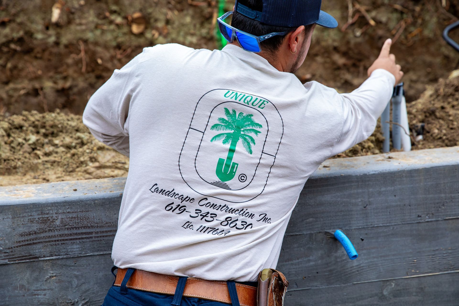 Man in tan shirt with company logo, pointing, standing by a wall.