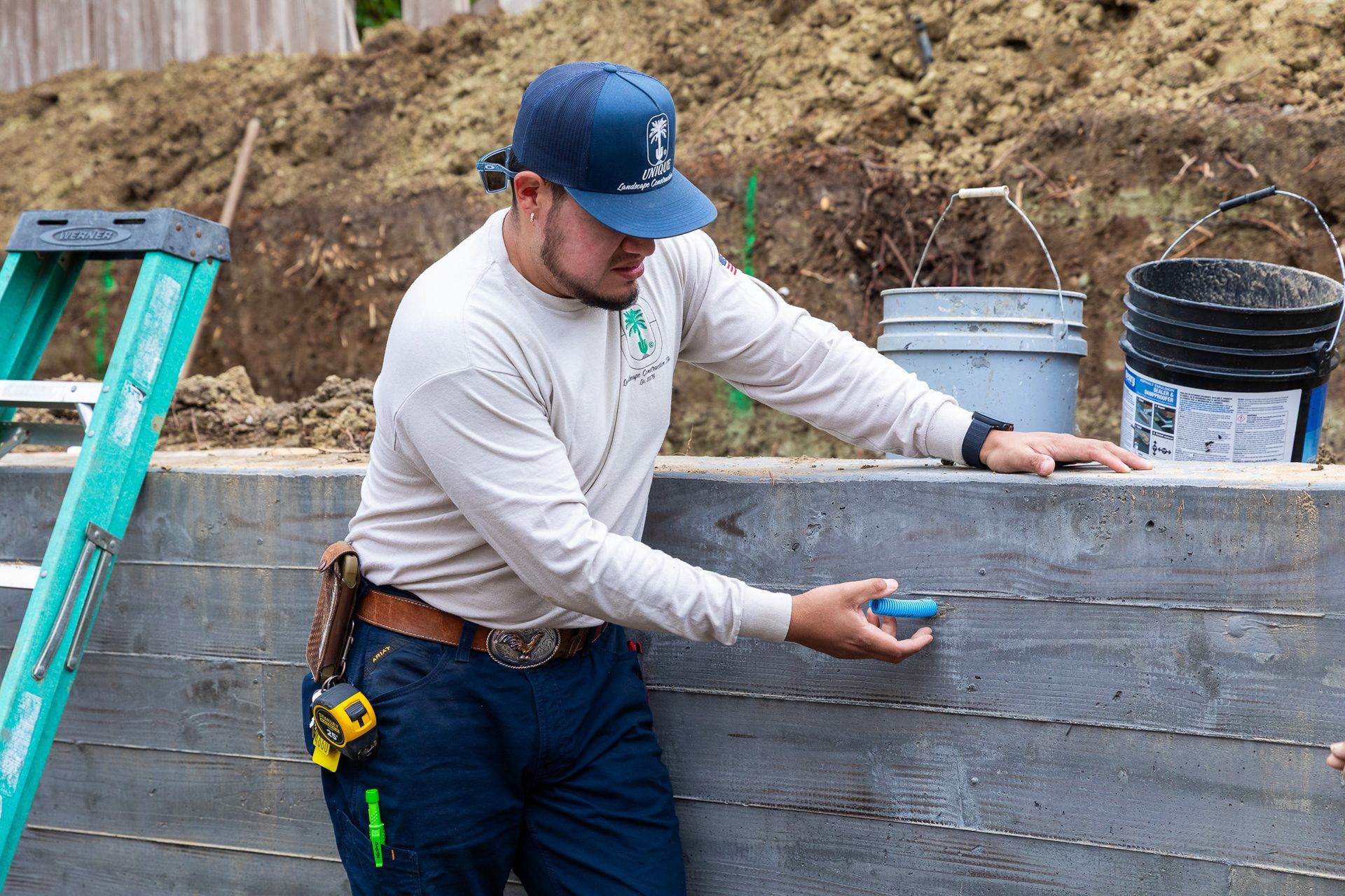 Man in cap and work clothes examines a blue pipe against a concrete wall.
