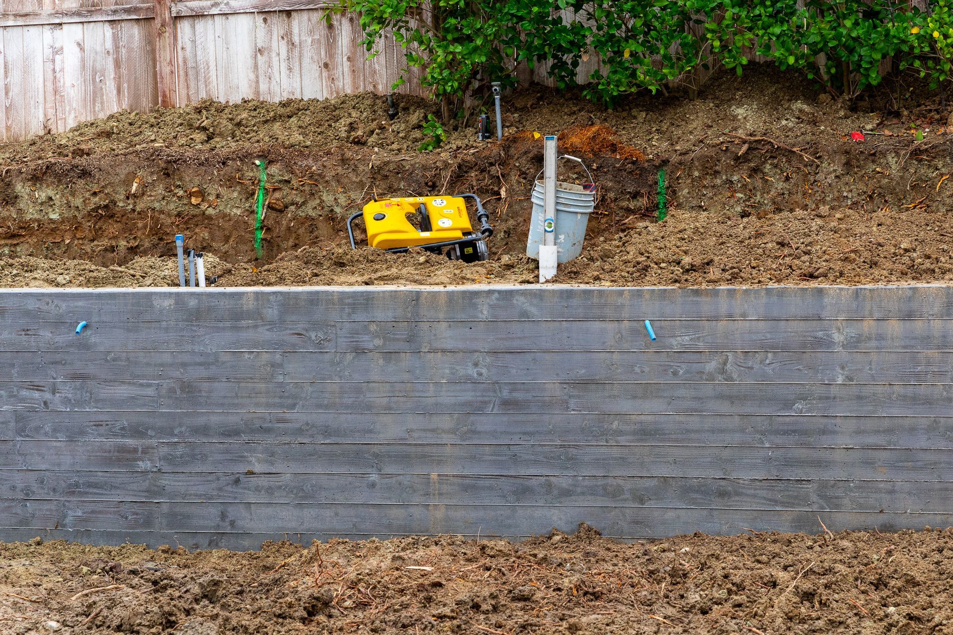 A retaining wall of grey wood-like concrete with dirt in front and a hillside behind with a yellow machine.