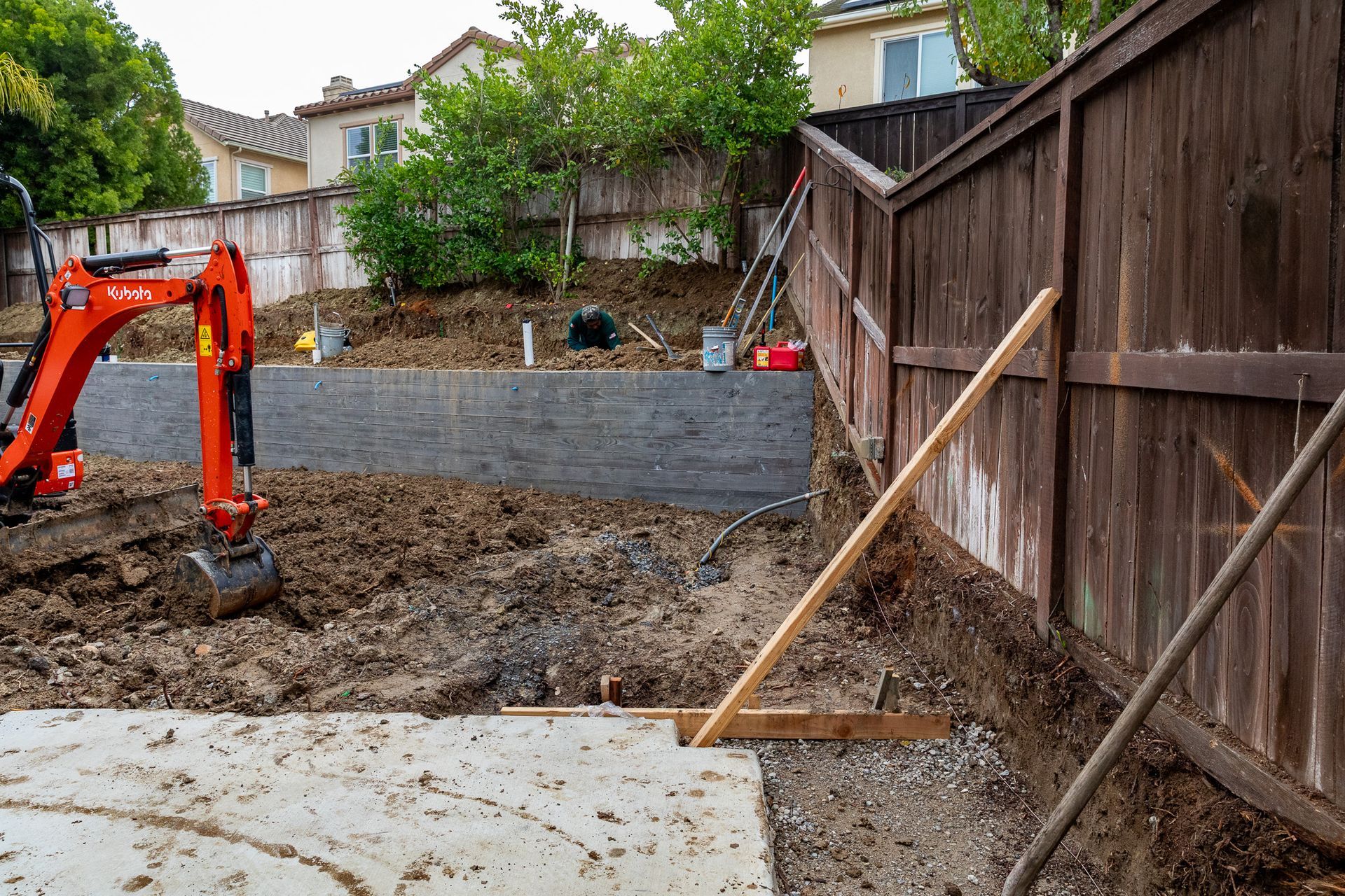 An orange excavator sits on a dirt lot next to a new grey retaining wall near a wooden fence in a suburban backyard.