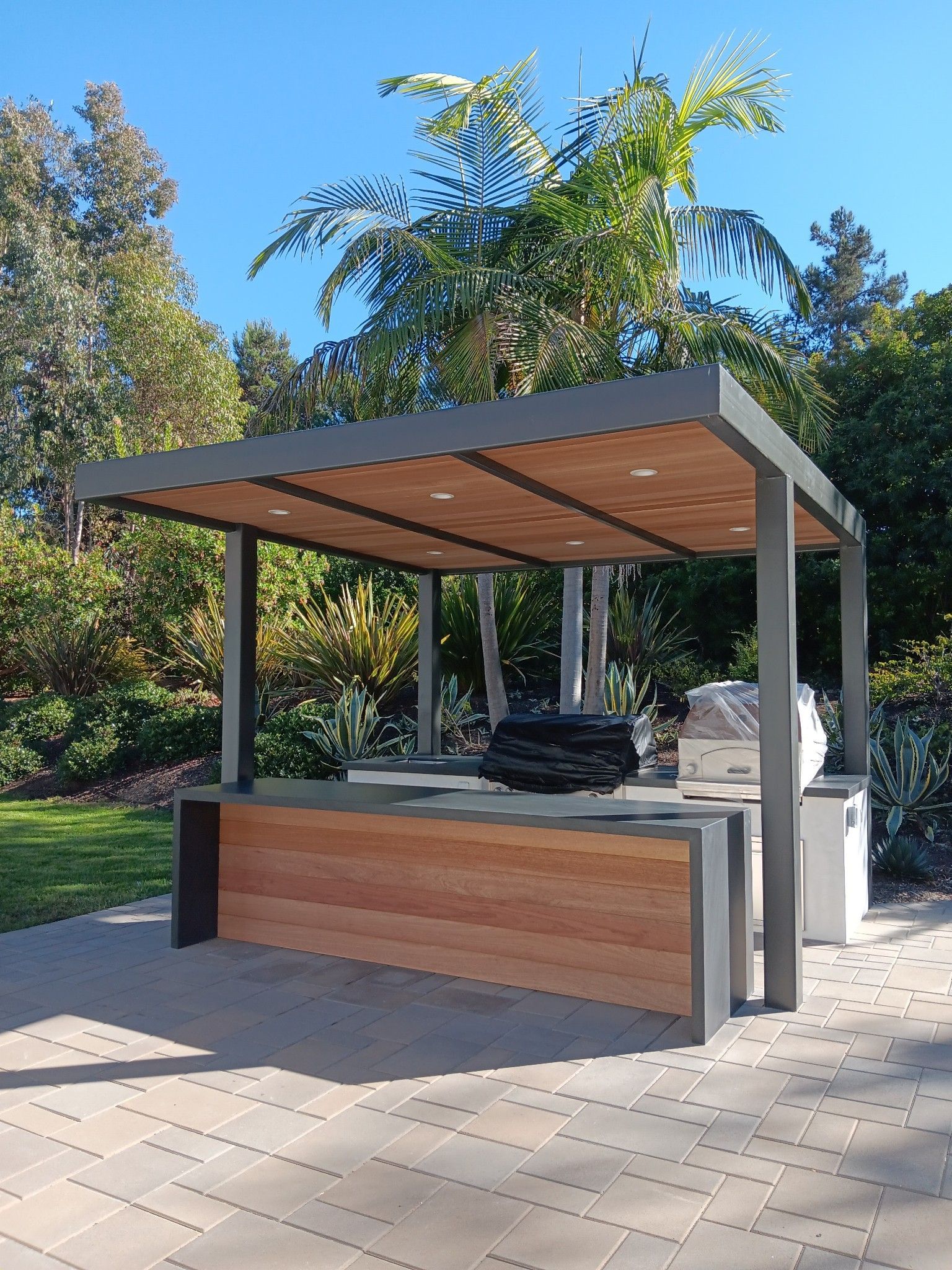 Outdoor kitchen with wood and gray metal construction; blue sky and greenery in the background.