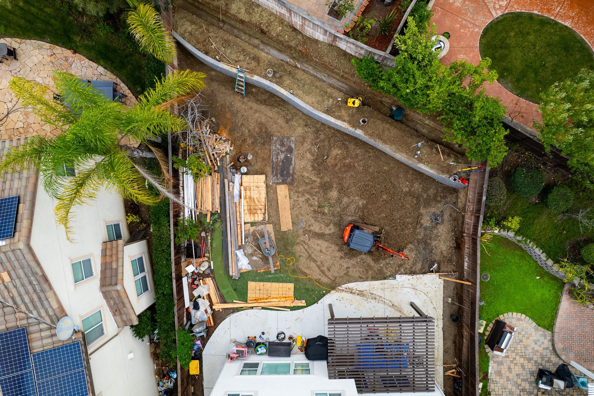 Aerial view of a residential backyard under construction with a small excavator, building materials, and a patio area.