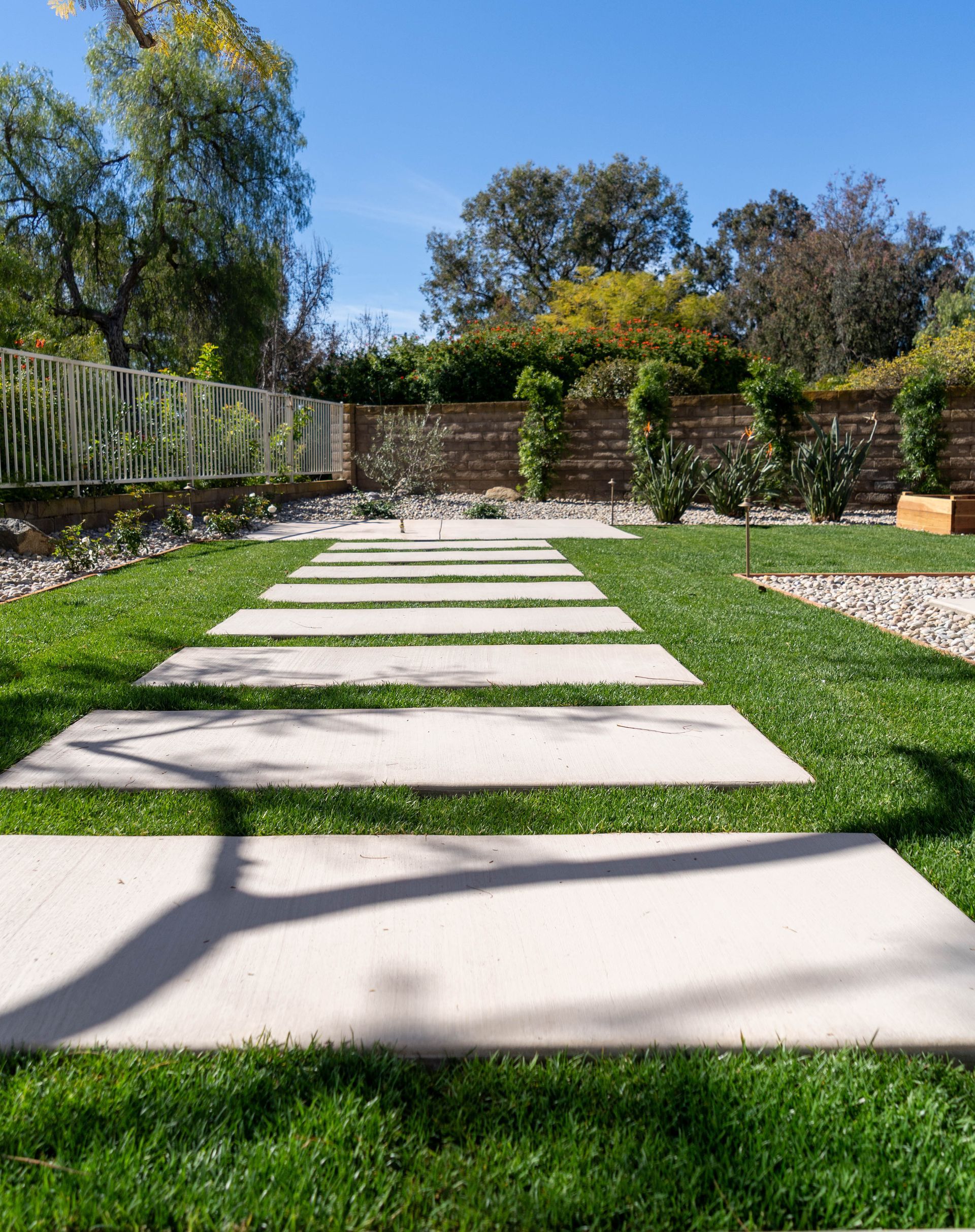 A stone pathway leads through a lush green lawn toward a wooden fence in a bright, sunlit backyard.