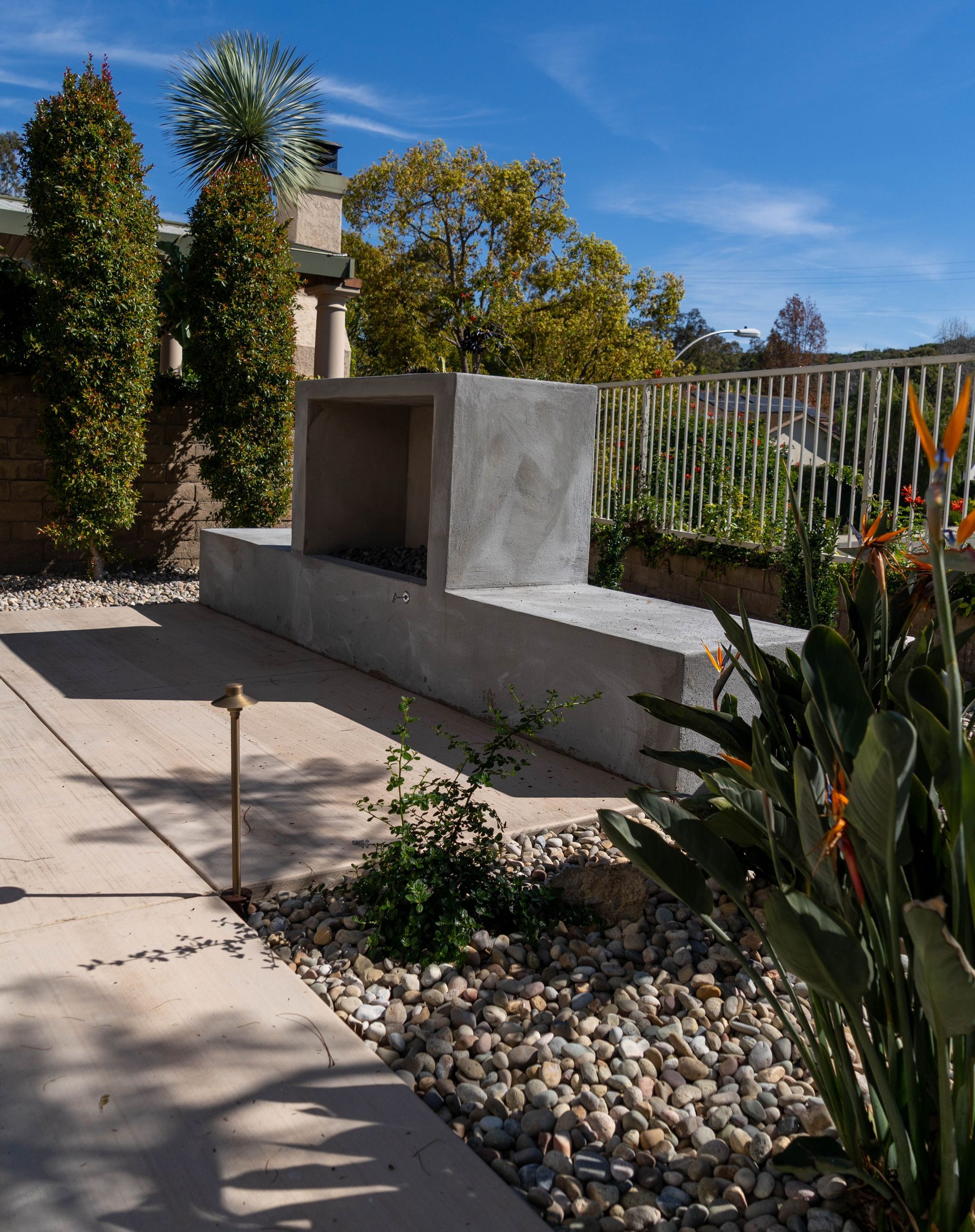 A minimalist concrete outdoor fireplace stands on a patio, surrounded by rocks, greenery, and a white fence under blue sky.