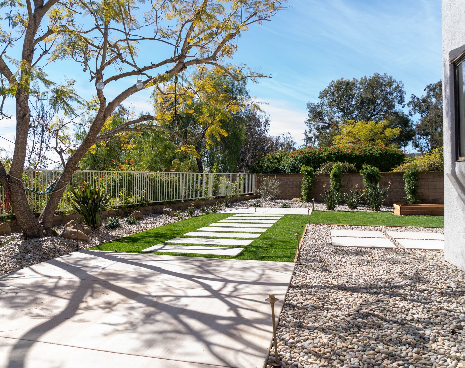 A backyard featuring a concrete patio, a stone walkway through green grass, surrounding gravel, and a large tree.