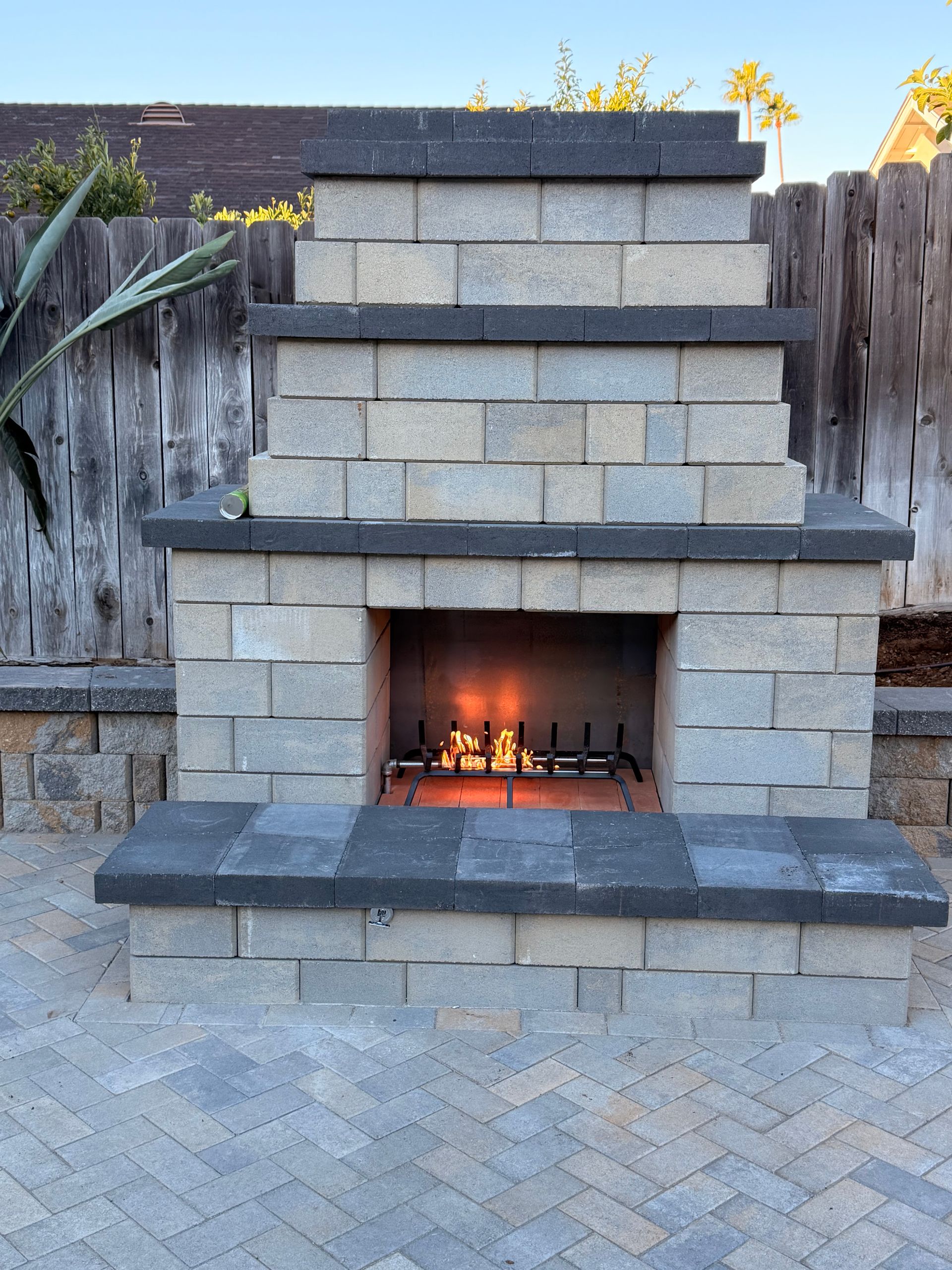 Stone outdoor fireplace with fire burning, set against a wooden fence and blue sky.