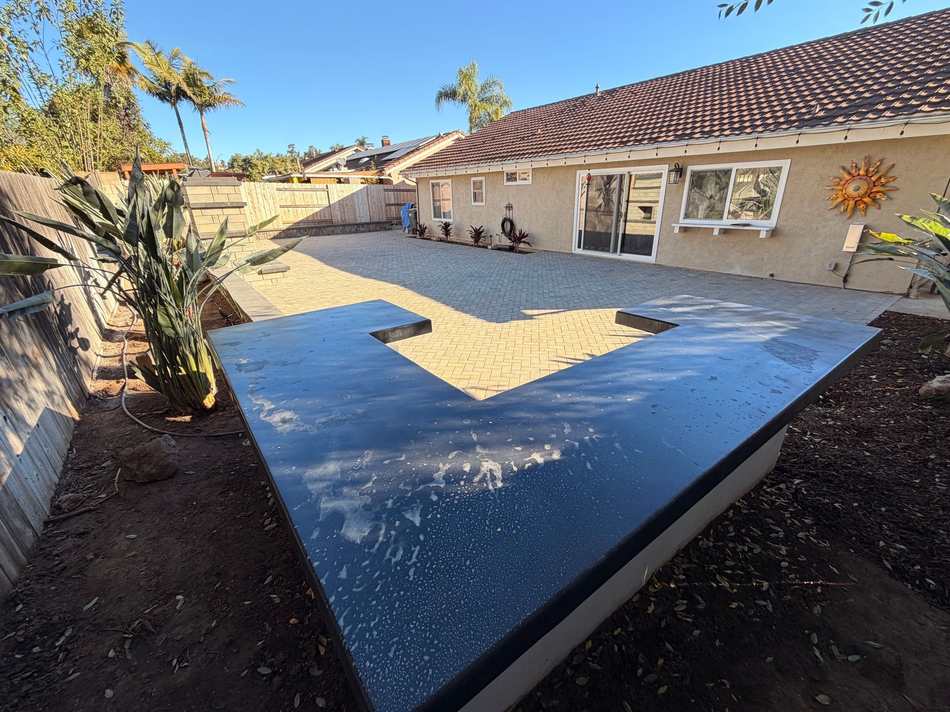 A backyard features a dark, L-shaped outdoor kitchen counter on a gravel patio beside a tan stucco house.