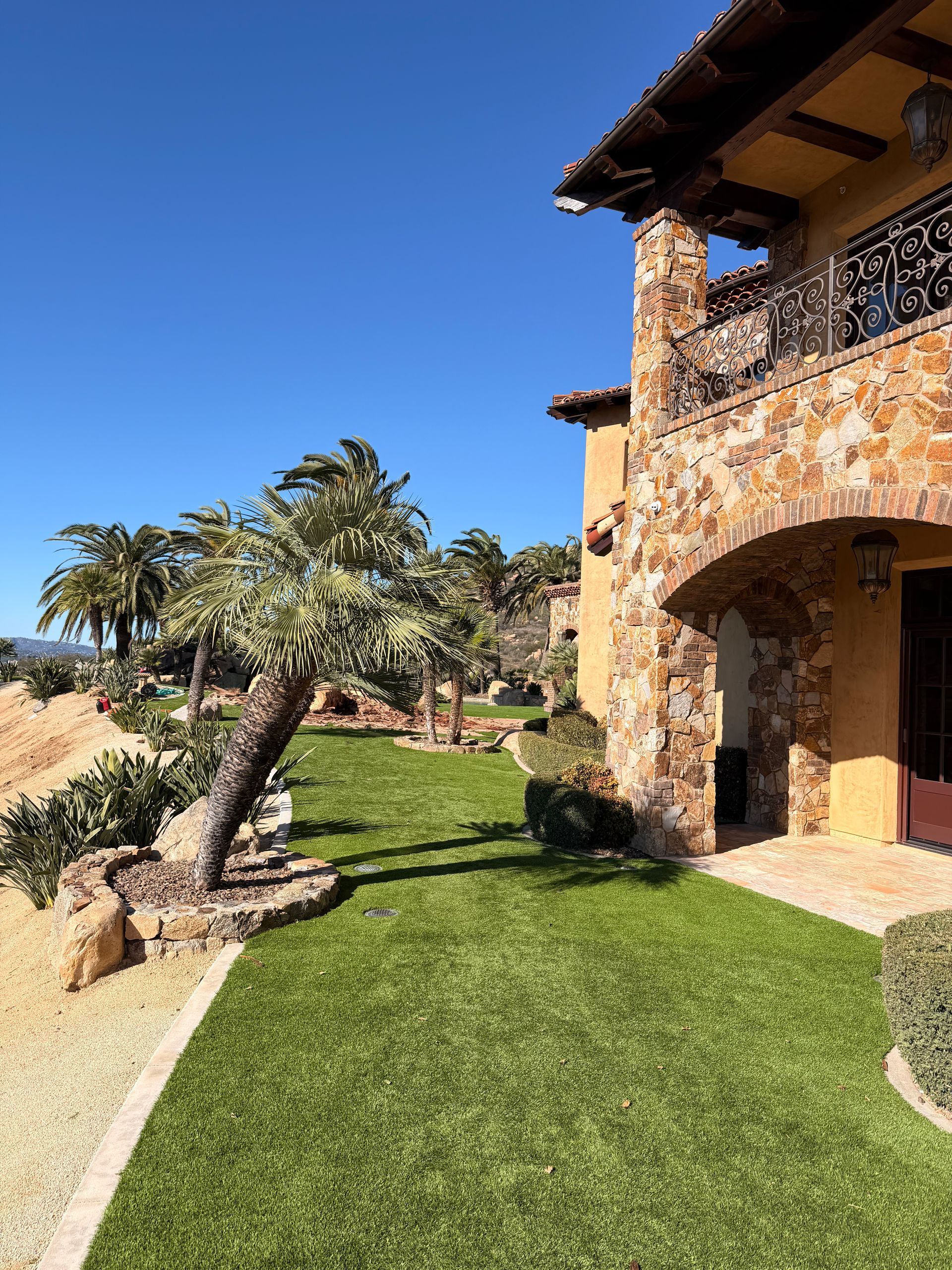 Two-story stone home with arched doorway and balcony, green lawn, palm trees, and blue sky.