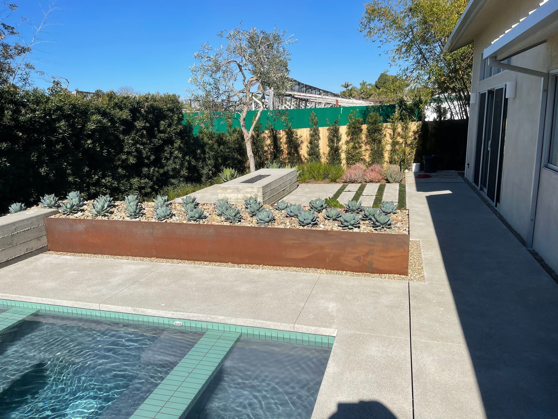 A rectangular, rusted metal planter filled with succulents sits next to a pool on a concrete patio in a sunny backyard.