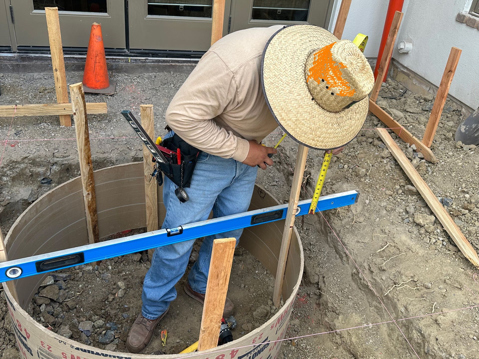 Construction worker in straw hat using a level and measuring tape inside a circular formwork.