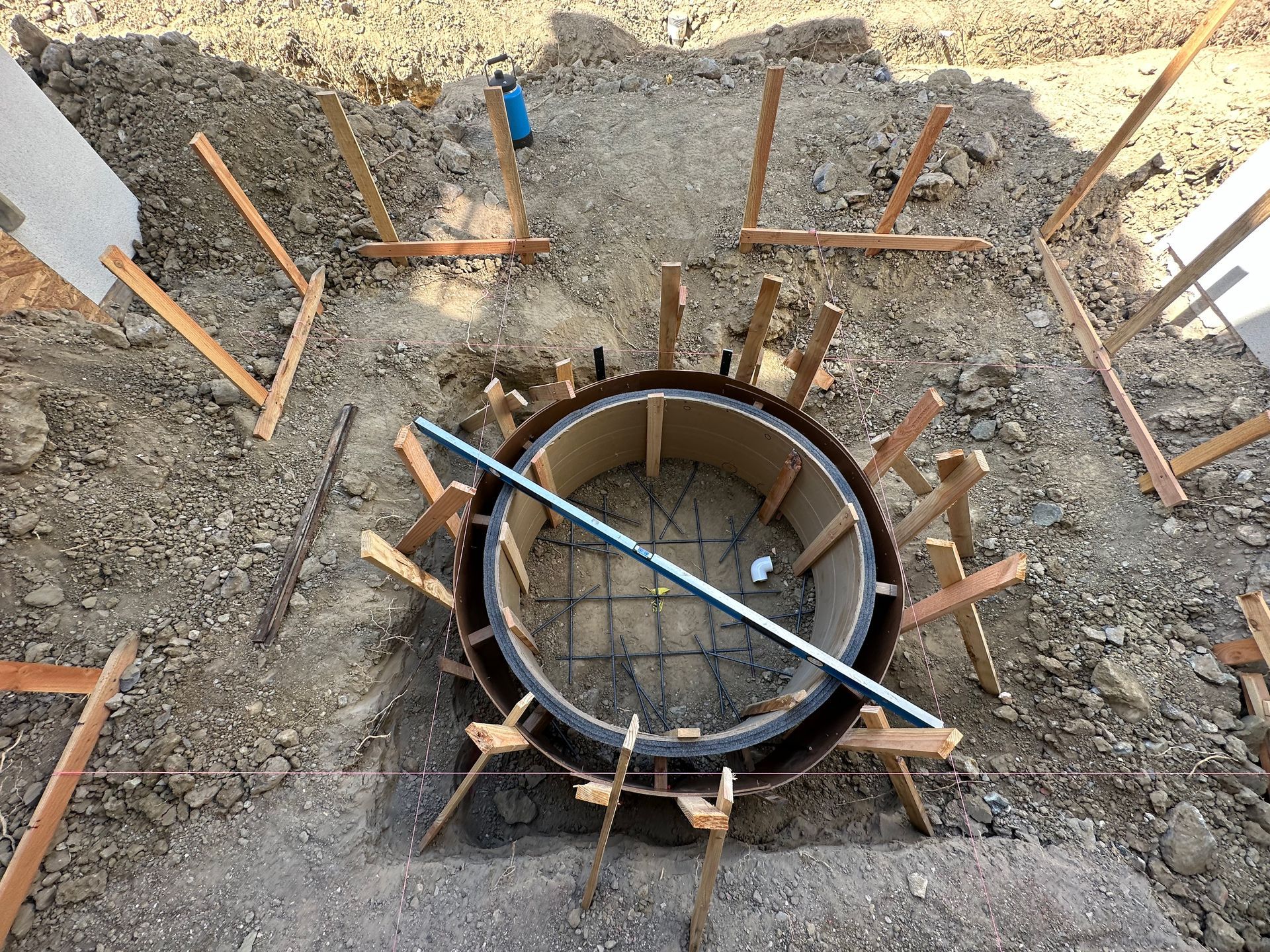 Circular formwork for concrete structure, surrounded by wooden supports and rebar grid in an earthen setting.