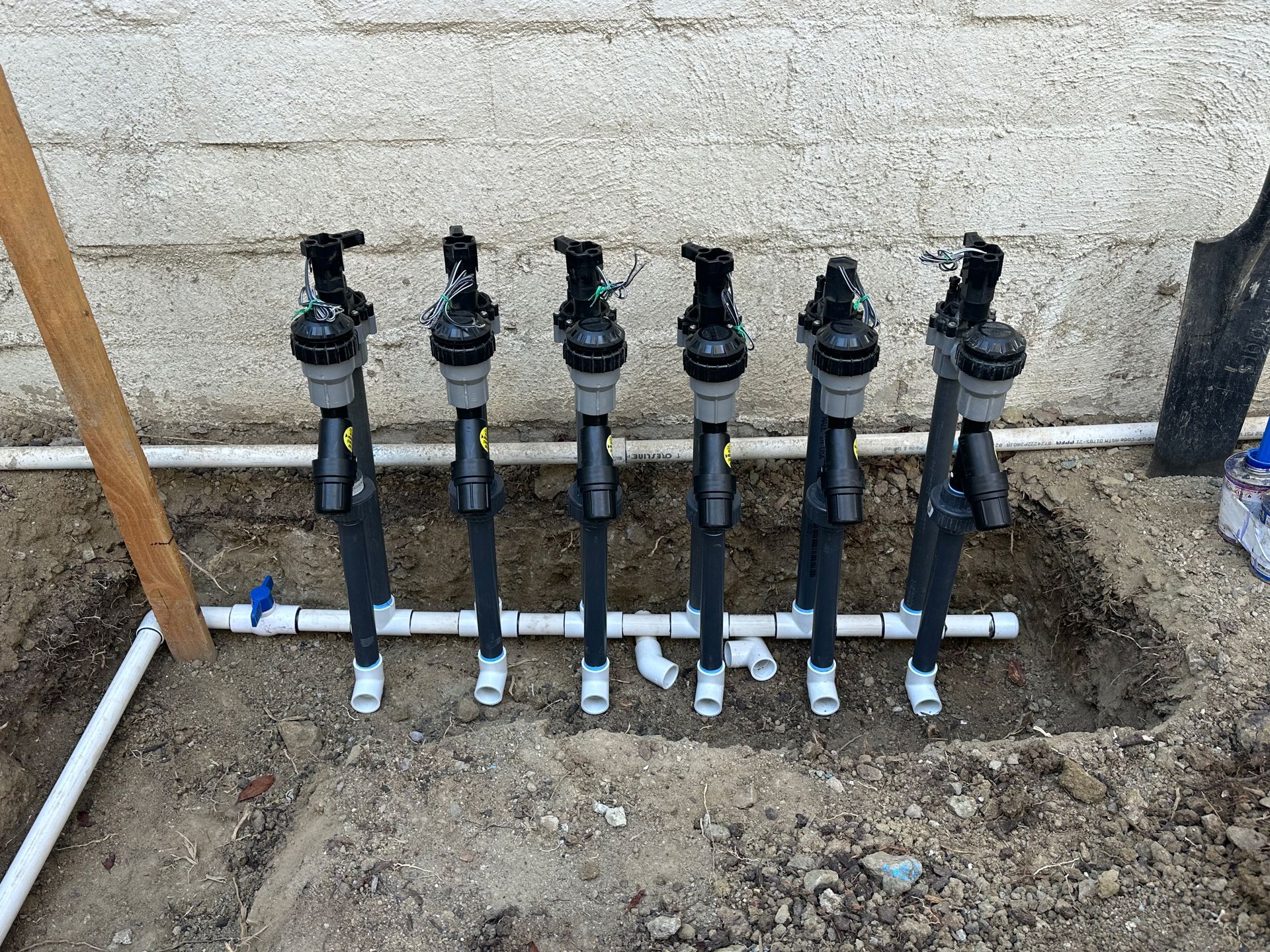 Six black sprinkler valves installed in a trench against a stucco wall, connected by white PVC pipes.