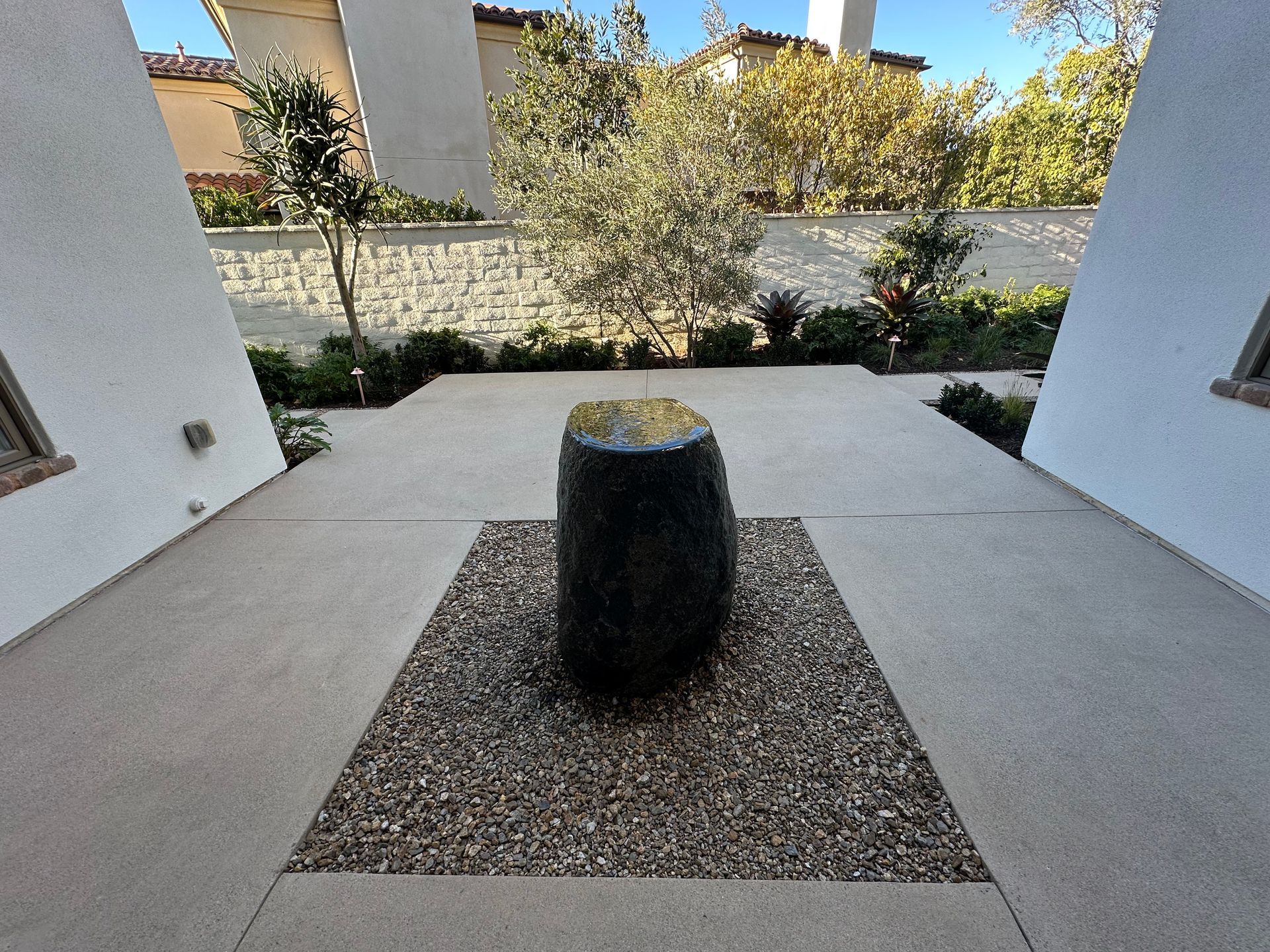 Stone water feature in gravel bed on a concrete patio, framed by white buildings and greenery.