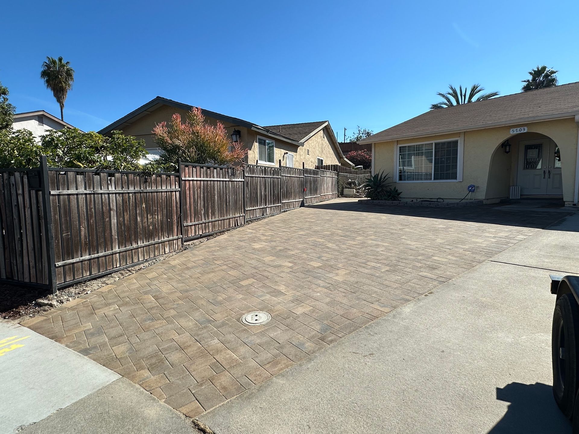 Brick paved driveway next to a weathered wooden fence and a one-story tan house on a sunny day.