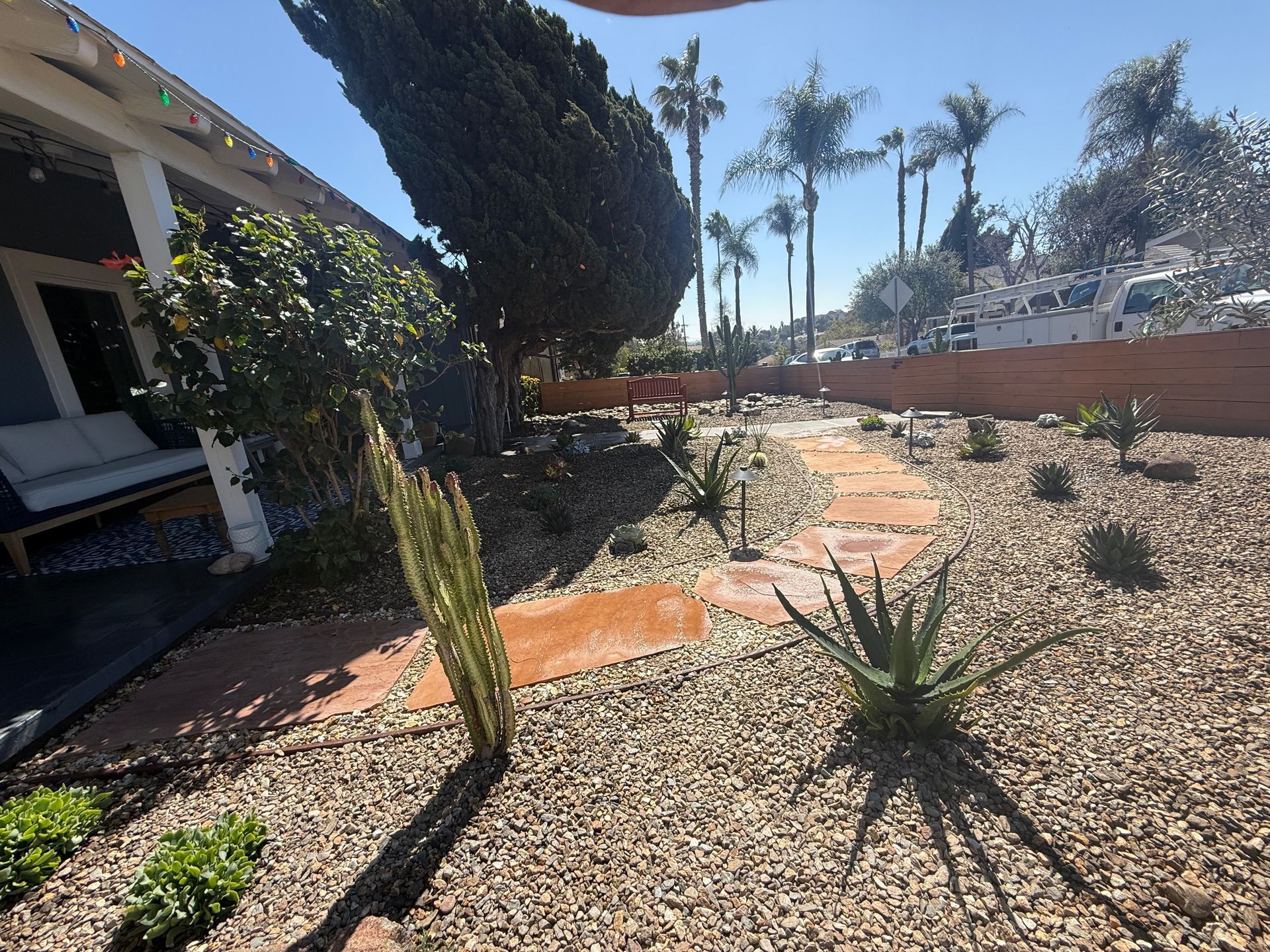 A sunny yard with a stone path, cacti, and gravel, next to a porch with a sofa.