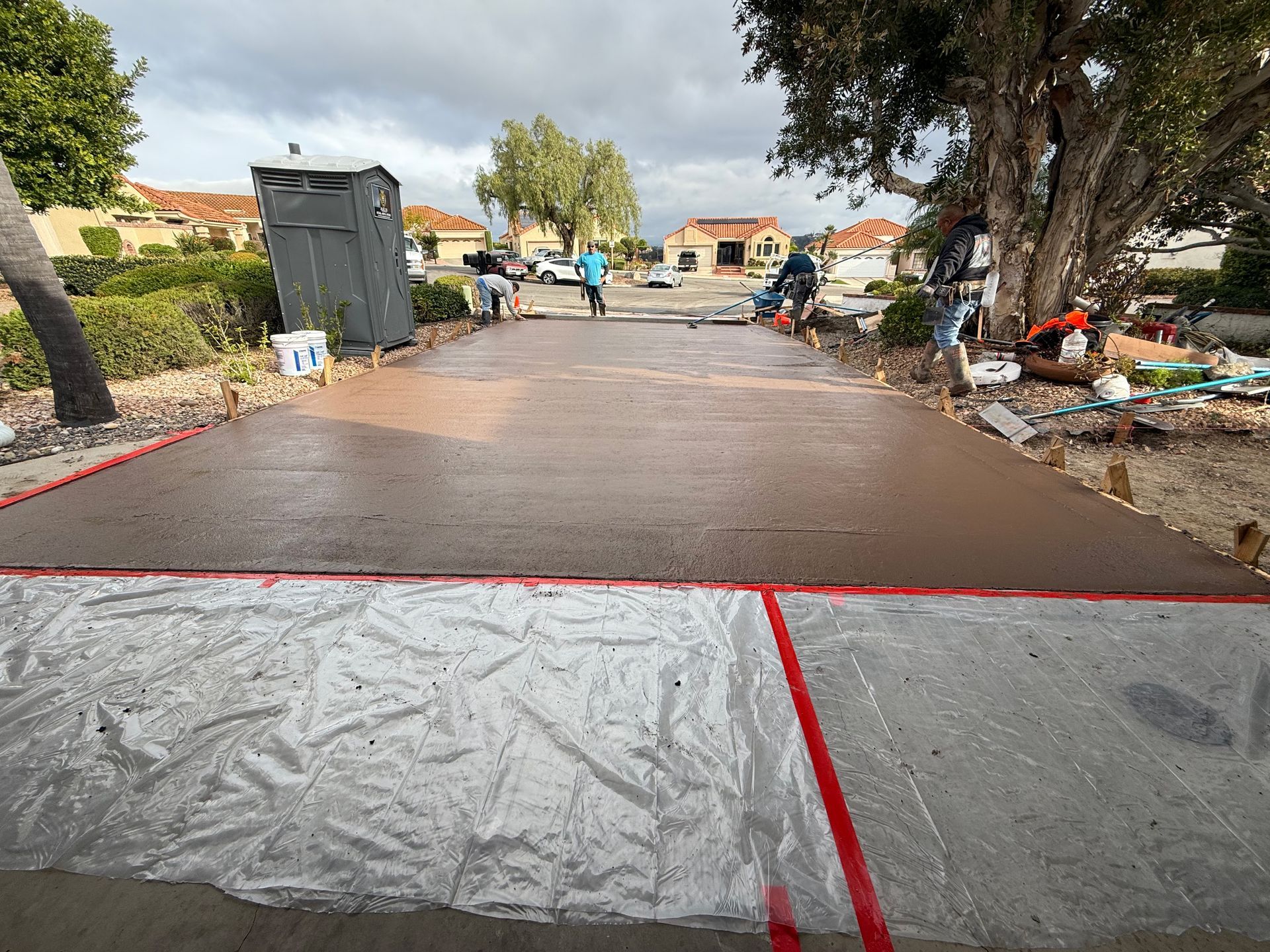 Concrete being poured on driveway; workers in blue shirts, plastic sheeting in foreground, houses in background.