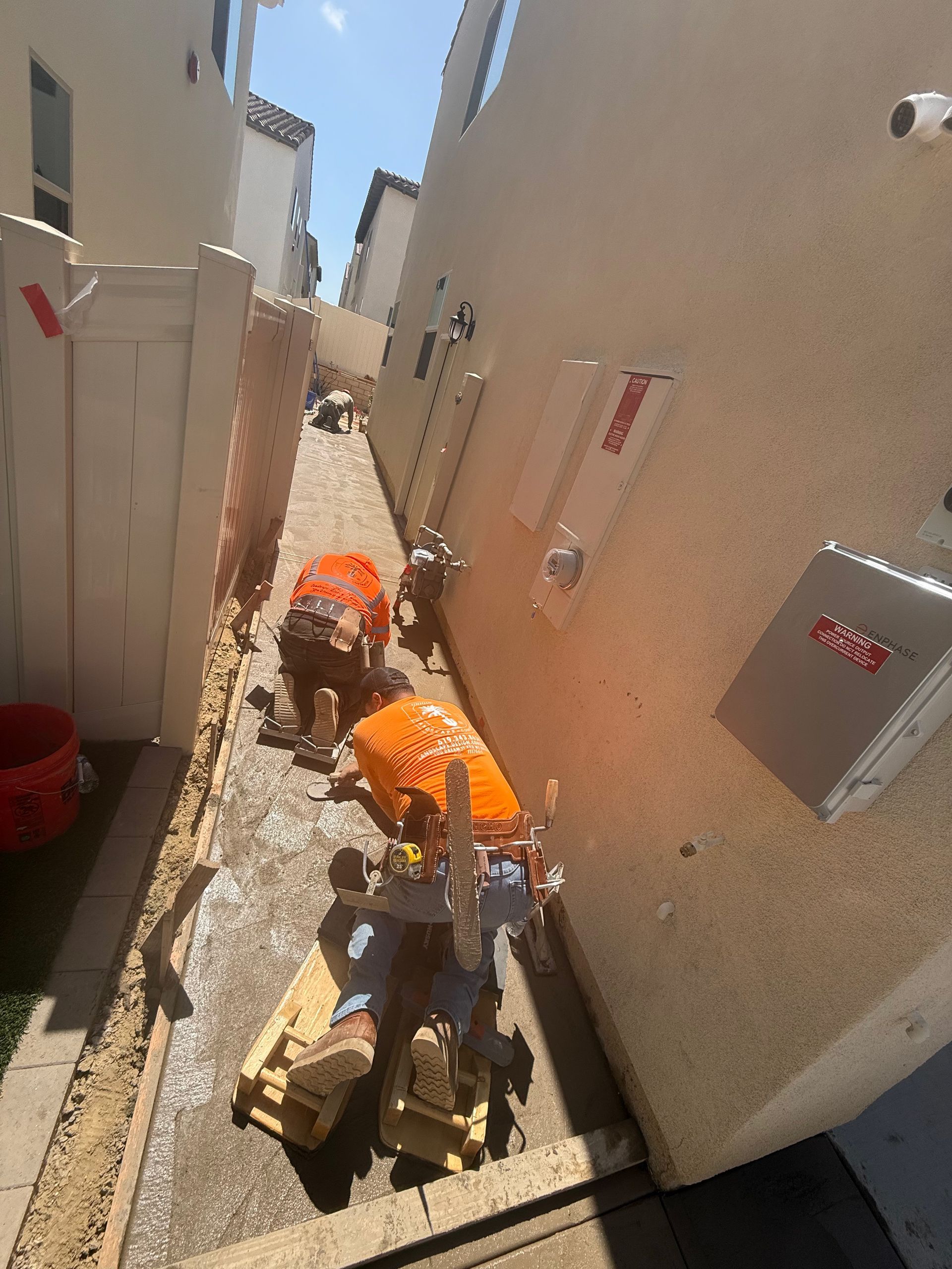 Construction workers in trench, orange vests, compacting soil, between buildings.