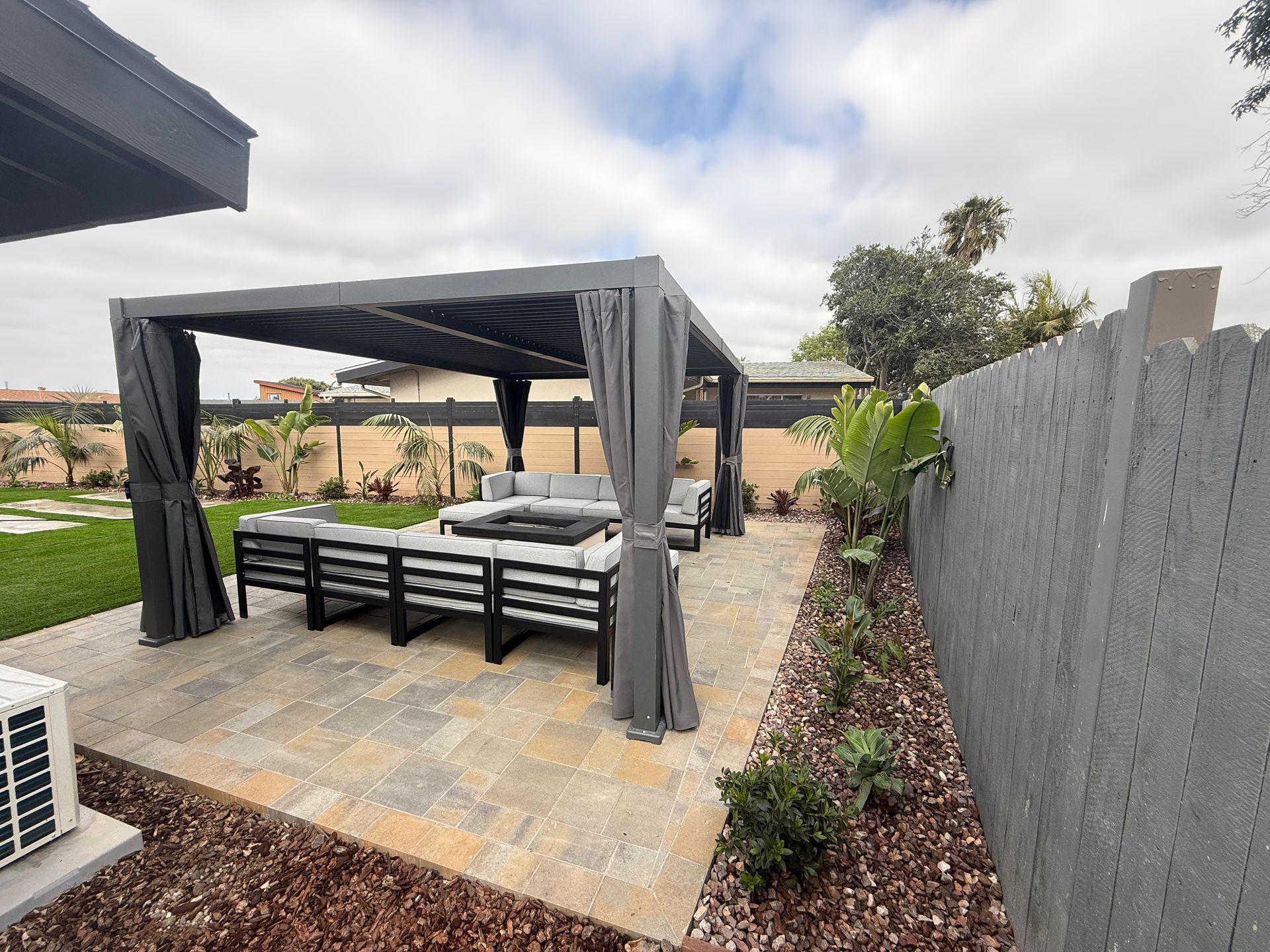 Patio with a pergola, seating, and a fire pit, near a gray fence and greenery under a cloudy sky.