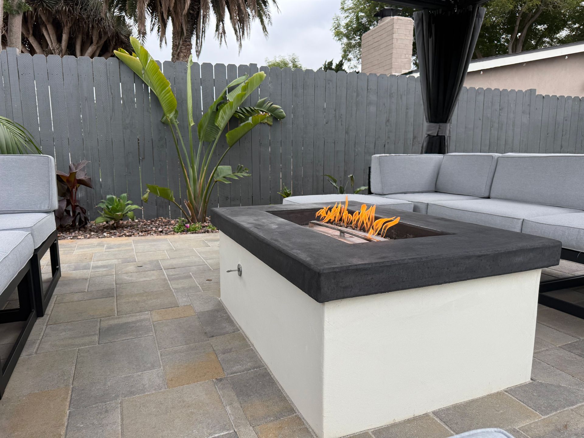 Outdoor seating area with lit fire pit. Gray and white furniture, gray fence, and tropical plants.