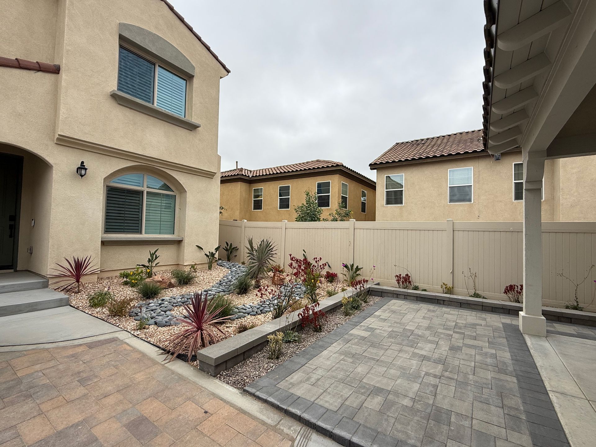 Two-story beige house with landscaped front yard, adjacent to a patio with brick pavers.