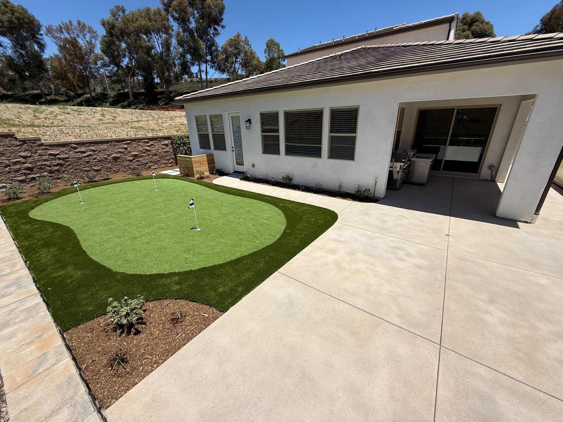 Backyard with putting green, patio, and white house under a sunny sky.