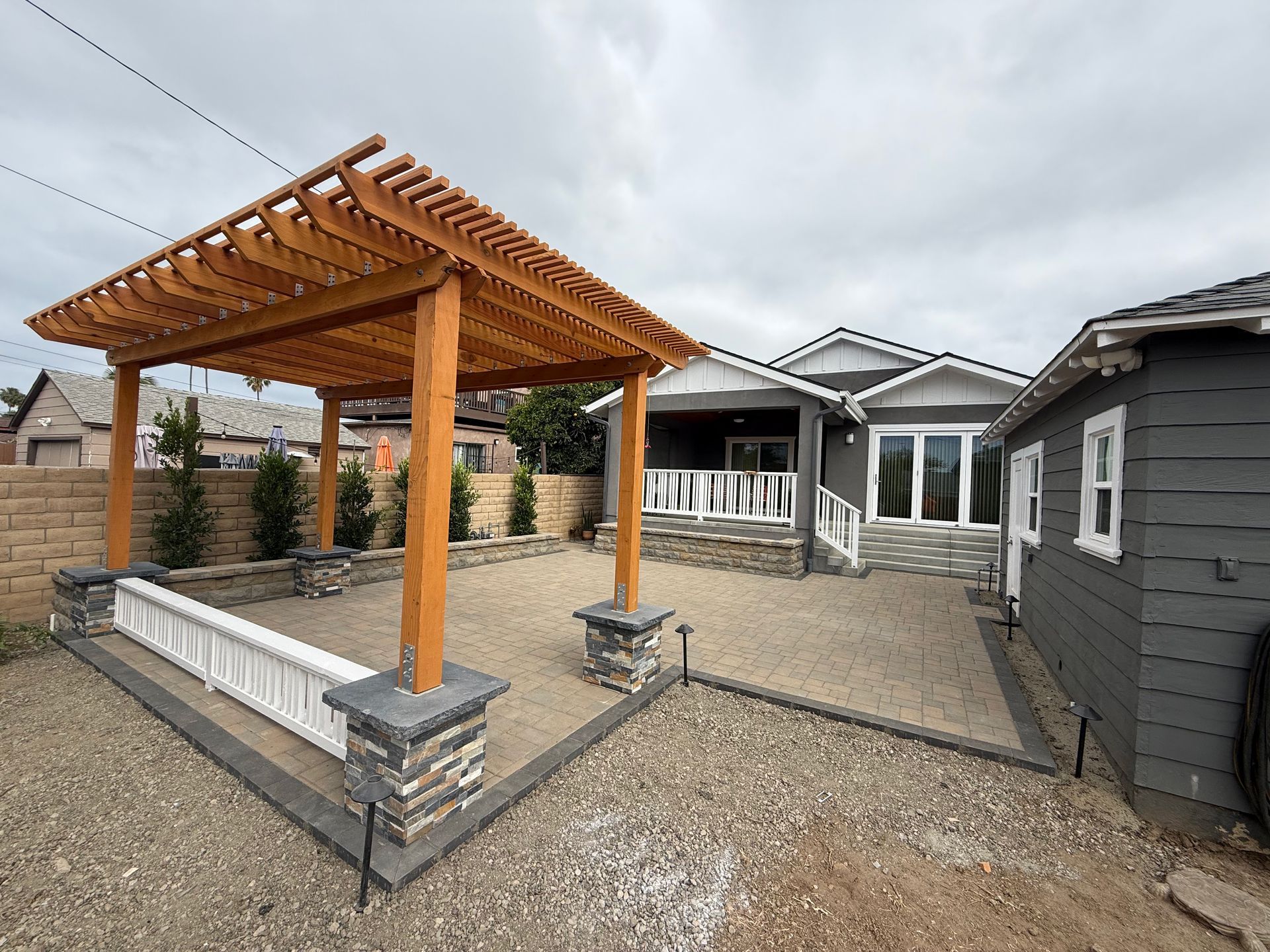 Wooden pergola over gravel patio; gray house and outbuilding in background, cloudy sky.