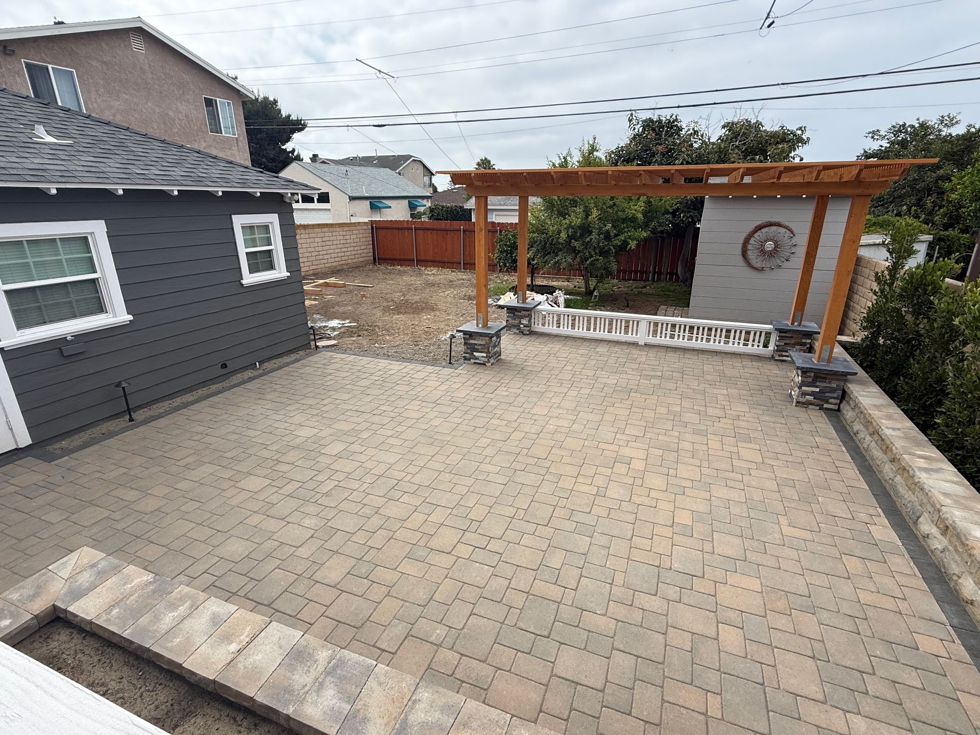 Patio with paved surface, pergola, and gray-sided shed. Backyard with a brown fence. Overcast sky.