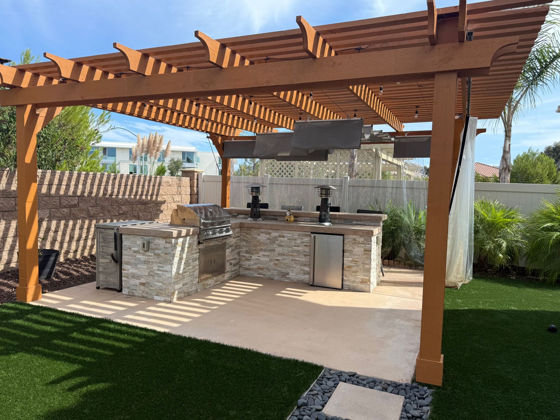 Outdoor kitchen with brown pergola over a stone-faced counter, grill, and appliances on concrete patio with green lawn.