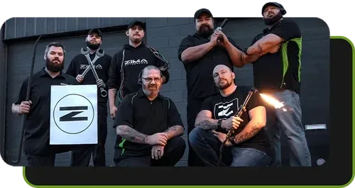 Zima team posing outdoors, holding a sign with a black Zima logo, one seated beside a small fire