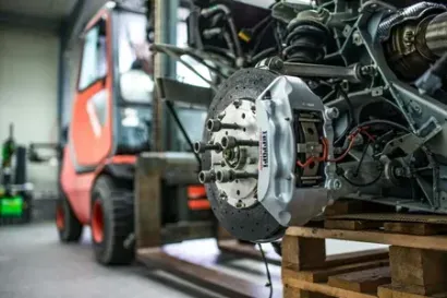 Forklift lifting a car chassis in a garage workshop, with exposed engine and brake assembly visible