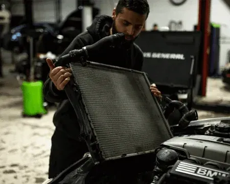 Mechanic inspecting a car radiator in a workshop, with tools and a BMW visible in the background