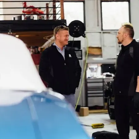 Max Zima and customer standing and talking in a workshop, with a blue car in the foreground.