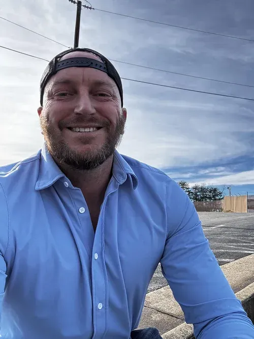 Smiling Max Zima in a light blue shirt and backward cap outdoors on a roadside under a cloudy sky
