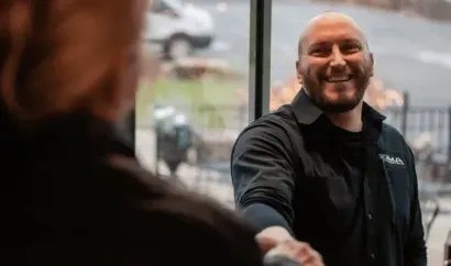Smiling Max Zima in a black jacket shaking hands indoors with a blurred person in the foreground