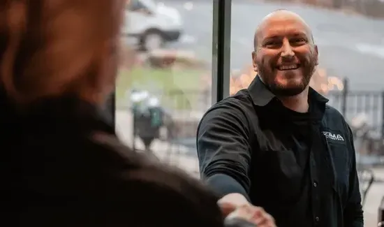 Smiling Max Zima in a black jacket shaking hands indoors with a blurred person in the foreground