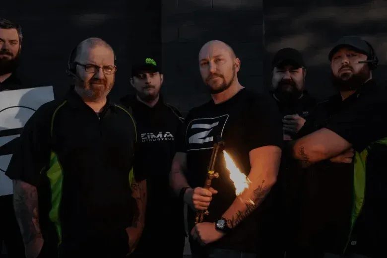 Zima team posing outdoors, holding a sign with a black Zima logo, one seated beside a small fire