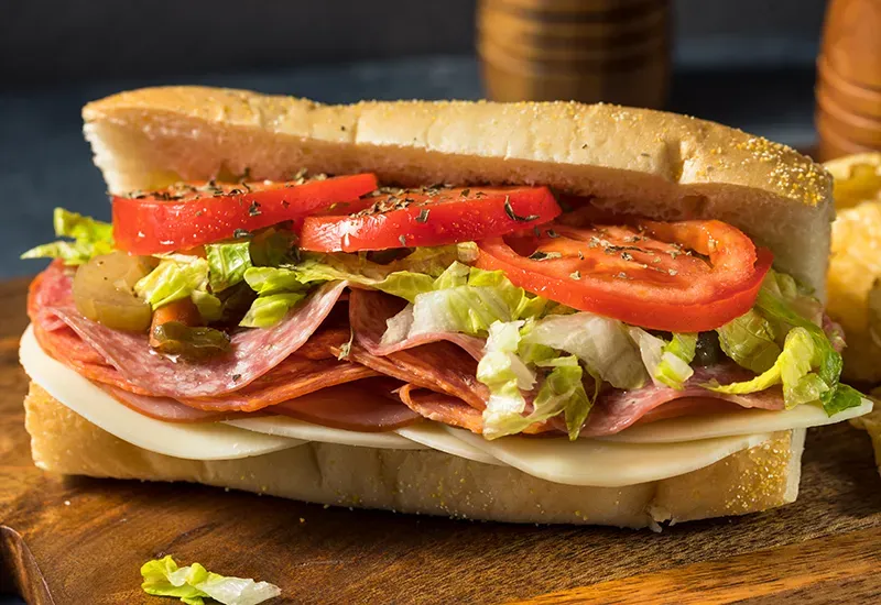 A sub sandwich with salami , cheese , lettuce and tomatoes on a wooden cutting board.