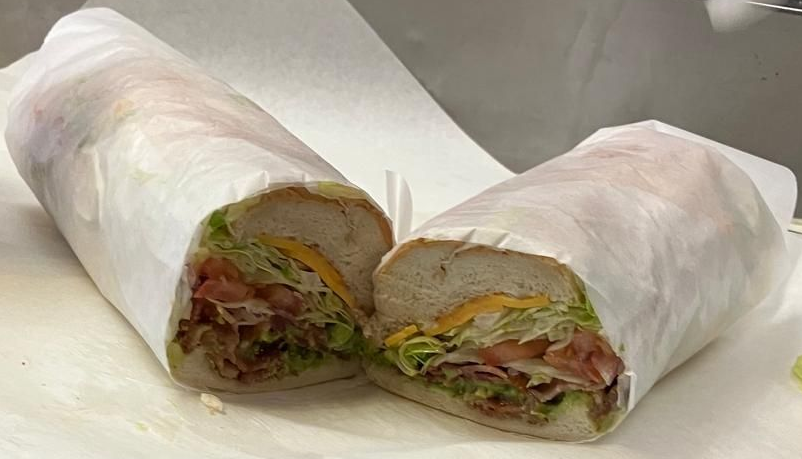 A sub sandwich is sitting on a wooden cutting board with vegetables.