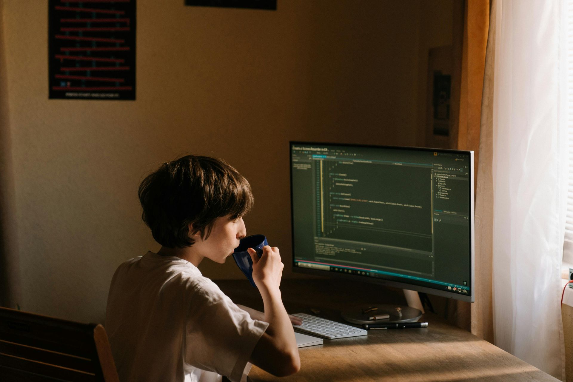 A young boy is drinking coffee while sitting at a desk in front of a computer monitor.