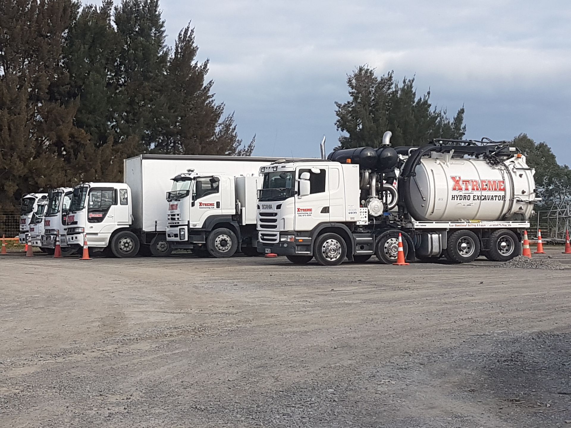 A row of trucks are parked in a gravel lot with trees in the background.